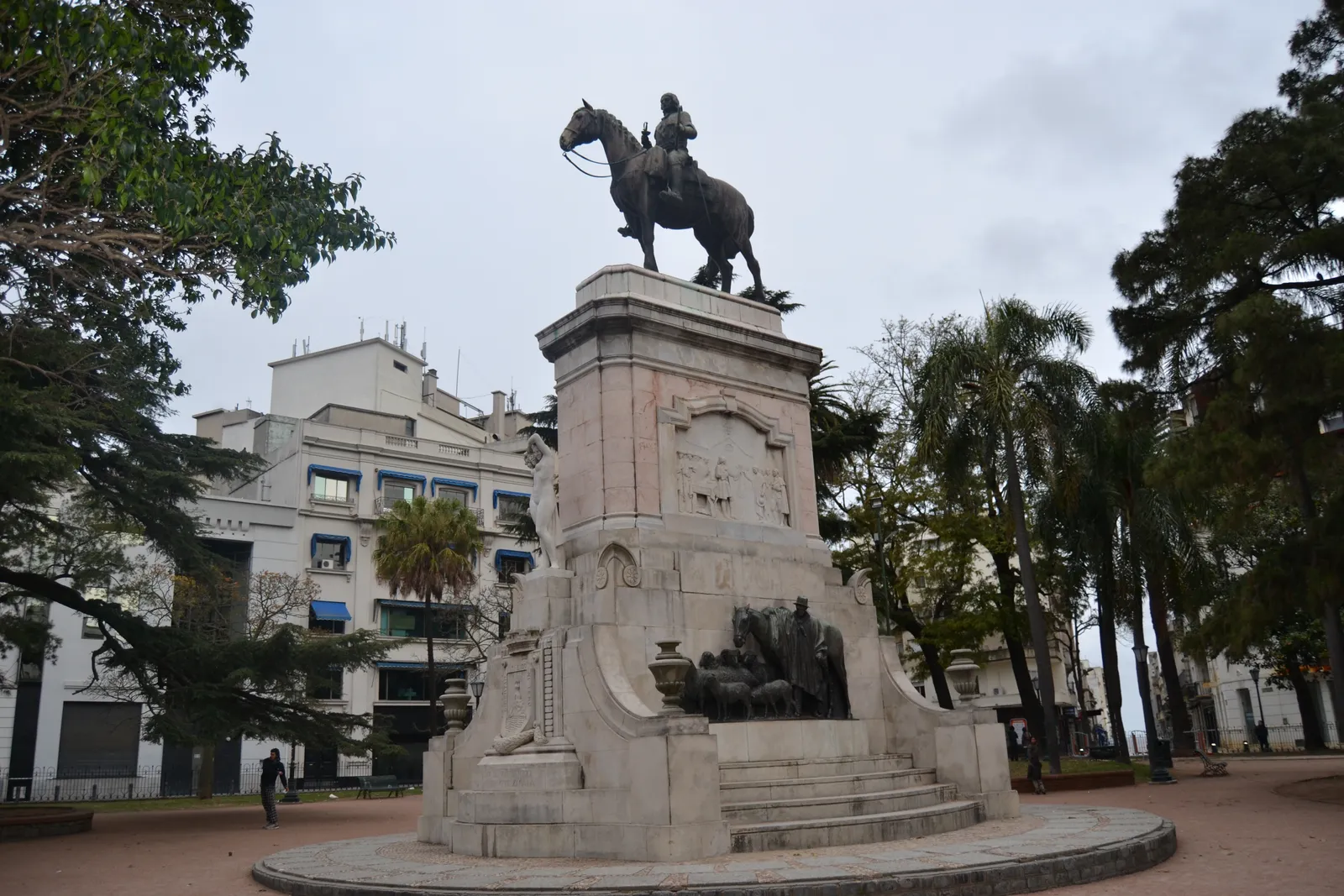 The equestrian statue of Bruno Mauricio de Zabala in Plaza Zabala, Montevideo, a bronze figure on horseback atop a tall stone pedestal with carved reliefs, surrounded by trees and old-city buildings
