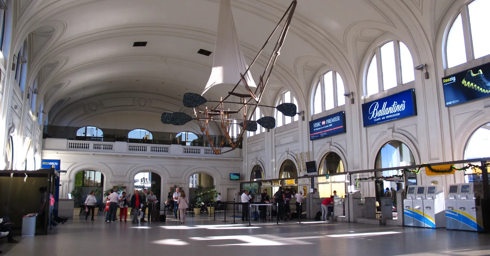 Inside Montevideo's ferry terminal, a vast vaulted hall from the early 20th century with arched windows and a mezzanine gallery, check-in counters and self-service kiosks below, and travelers with luggage crossing the floor