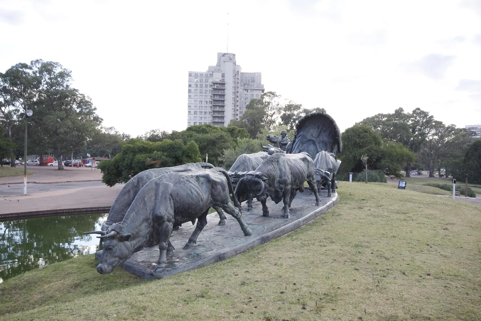 The La Carreta monument in Montevideo's Parque Batlle, a bronze team of oxen straining uphill on a grass bank to pull a covered wooden wagon with a driver on top, a pond in front and park trees behind