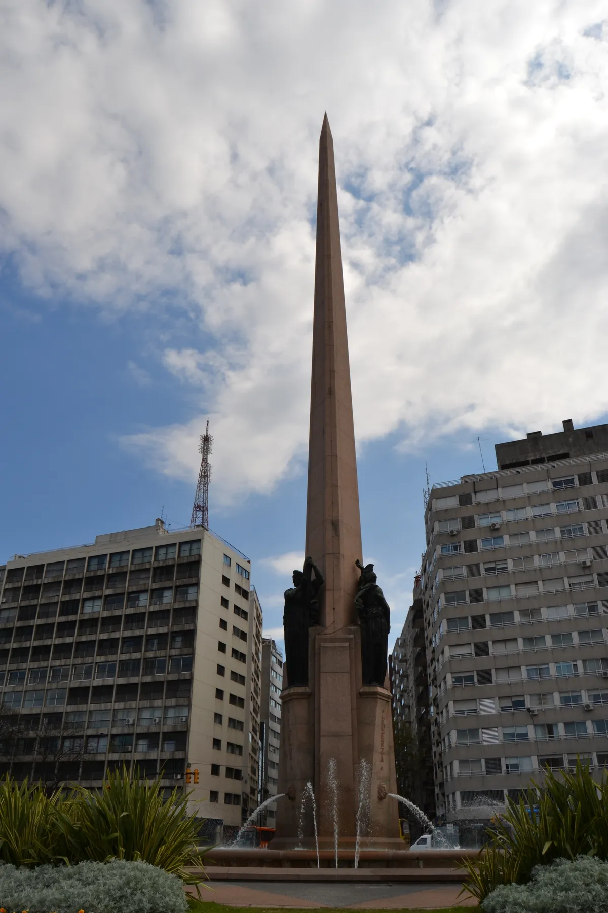 The tapering pink granite obelisk to Uruguay's 1830 constituent assembly in Montevideo, with three dark bronze figures standing against its base and fountains playing in front of it, apartment blocks on either side