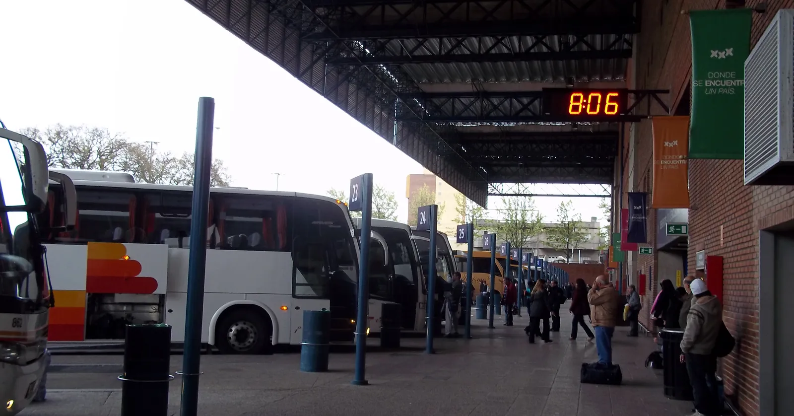 A row of long-distance coaches standing at the platforms of Montevideo's Tres Cruces terminal under a steel canopy, with a digital clock overhead and passengers waiting with luggage along the walkway