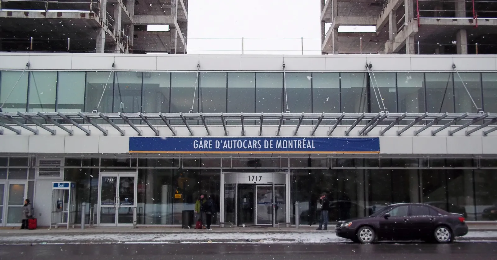 The glass entrance canopy of the Gare d'autocars de Montréal on rue Berri with the terminal's name on a blue sign above the doors and travellers wheeling luggage past in the snow