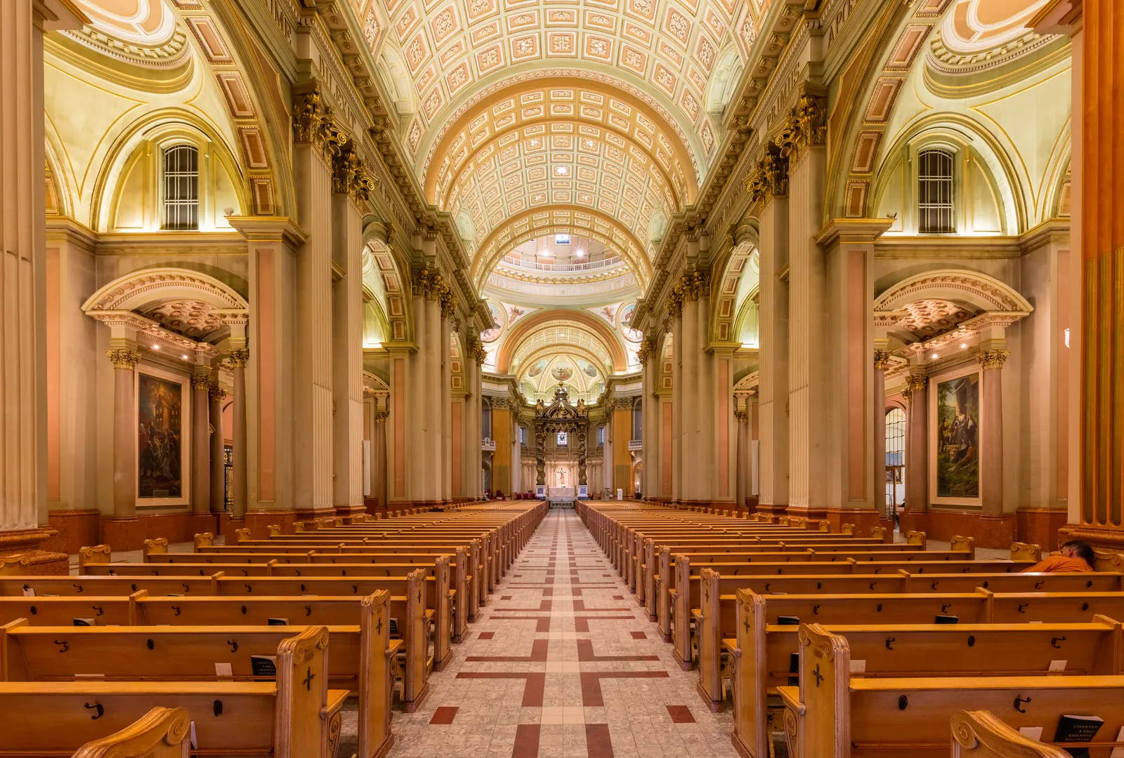 The nave of Mary, Queen of the World Cathedral in Montreal looking towards the canopied altar, with a coffered barrel vault overhead and rows of wooden pews on a patterned stone floor
