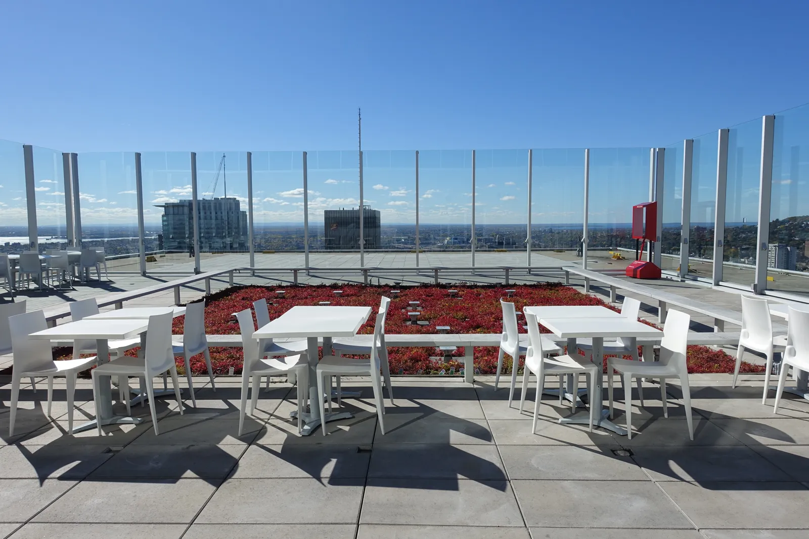 The open-air terrace at the top of Place Ville Marie in Montreal, white tables and chairs behind a glass balustrade with planted beds, downtown rooftops and the river spread out beyond