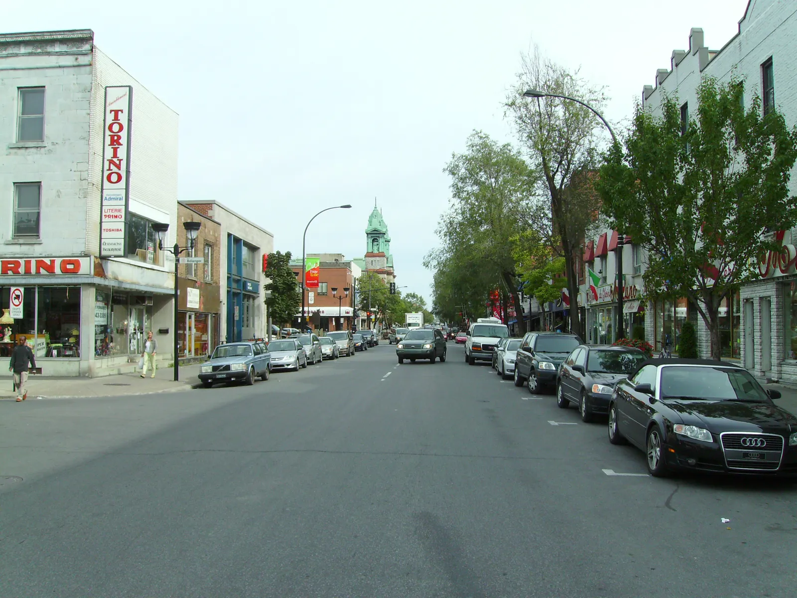 Boulevard Saint-Laurent running through Little Italy in Montreal, low brick shopfronts and cafés along both kerbs with a green church steeple rising further up the street