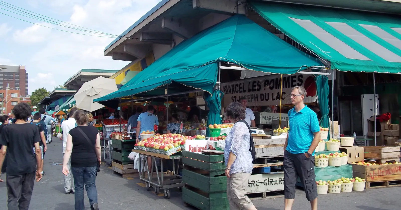Produce stalls under green awnings at Jean-Talon Market in Montreal, wooden crates of apples and tomatoes stacked along the aisle with shoppers walking between them