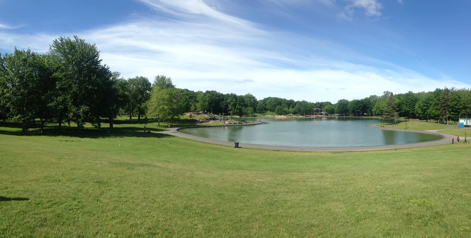 Beaver Lake on Mount Royal in Montreal on a clear summer day, a shallow oval basin ringed by mown grass and mature trees under a blue sky