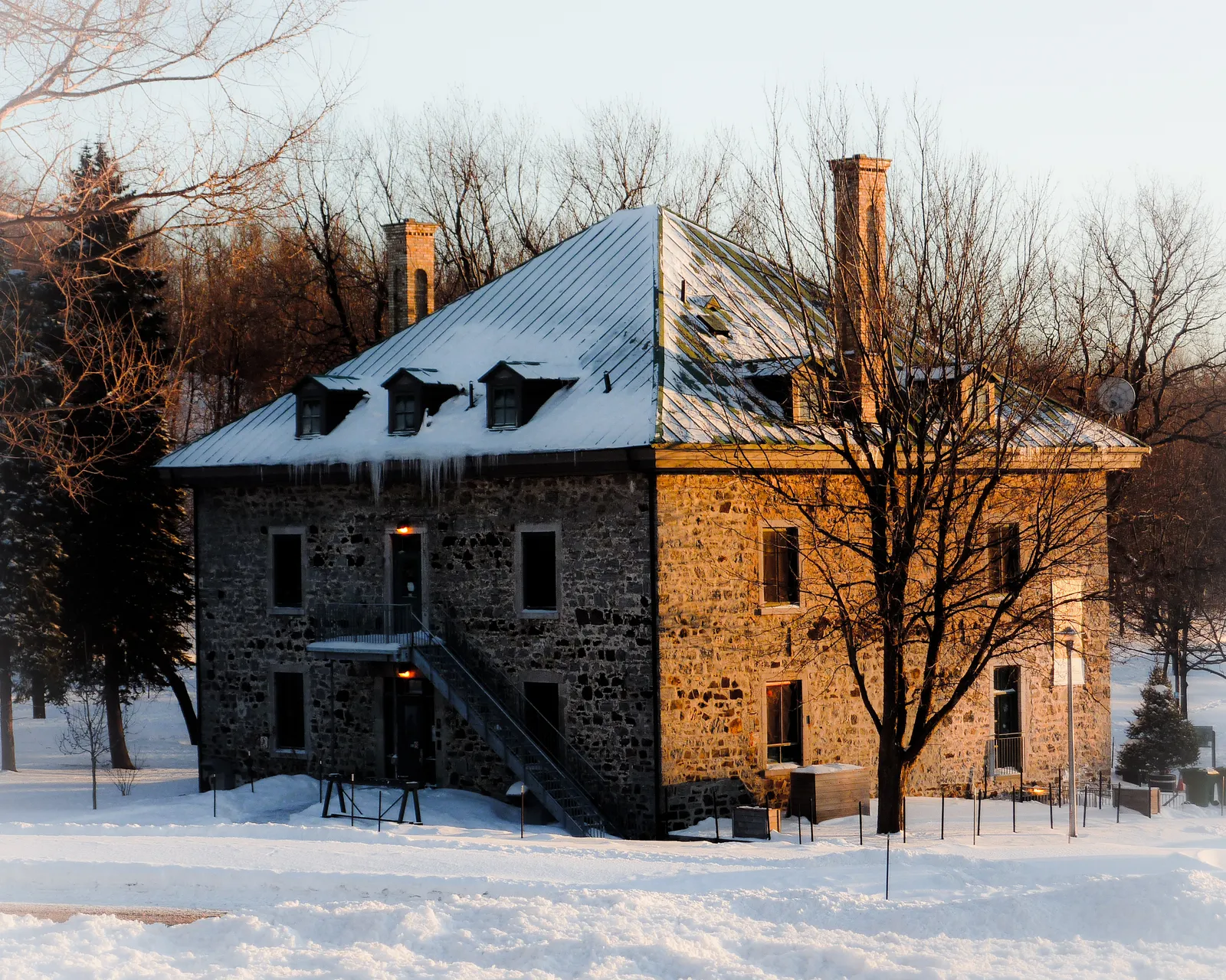 Smith House on Mount Royal in Montreal, a square two-storey fieldstone farmhouse with a pitched metal roof and two chimneys, standing in snow among bare trees