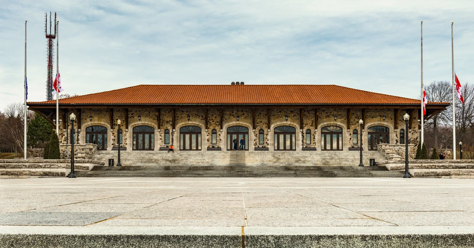 The front of the Mount Royal Chalet in Montreal, a long single-storey pavilion in rough stone with a red tiled roof, arched doorways along its face and flagpoles on the terrace in front