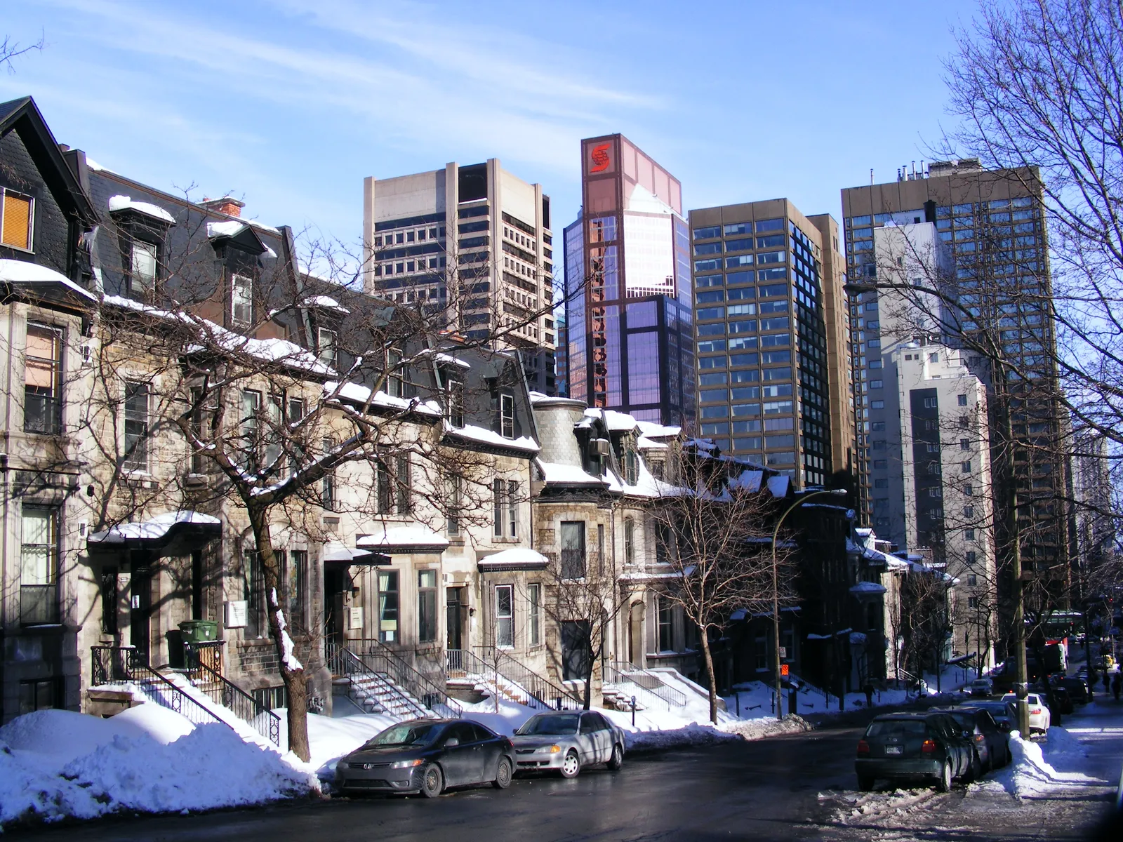 A terrace of Victorian greystone houses with outdoor staircases on Peel Street in Montreal's Golden Square Mile, snow on the pavement and modern downtown office towers rising directly behind them