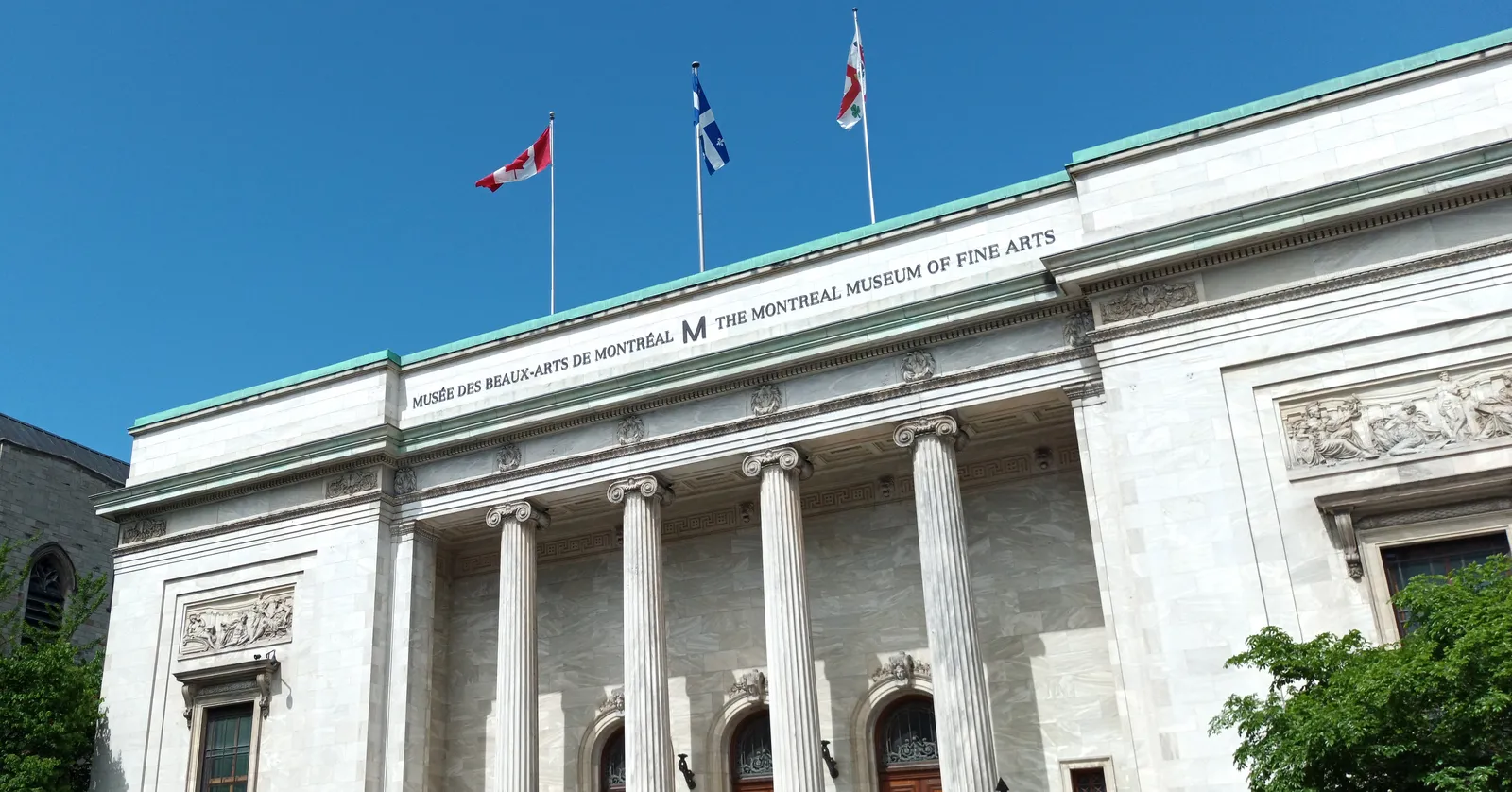 The white marble front of the Montreal Museum of Fine Arts on Sherbrooke Street, a row of tall columns under a frieze carrying the museum's name, with flags flying above it against a blue sky