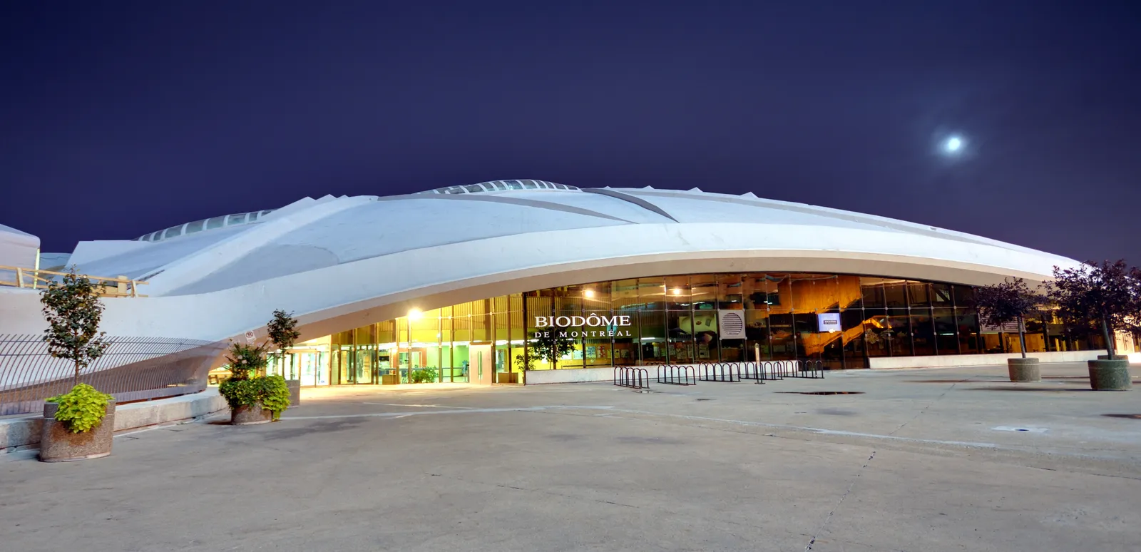 The Montreal Biodome at dusk, its long curved white roof lit from below with the building's name above the glazed entrance doors