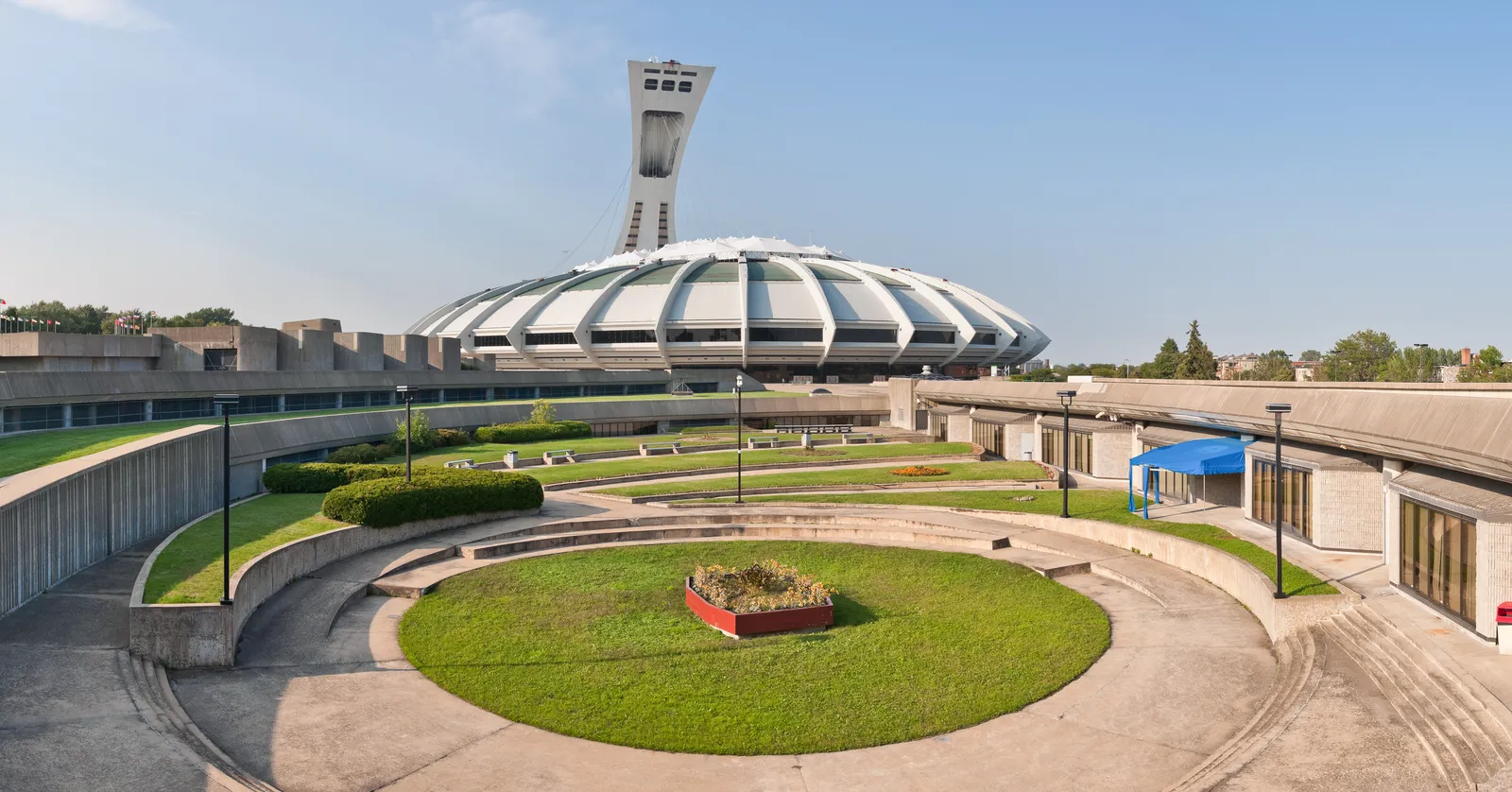 The ribbed white roof of Montreal's Olympic Stadium with the leaning concrete mast of the Montreal Tower rising behind it, seen across the empty circular plaza in front