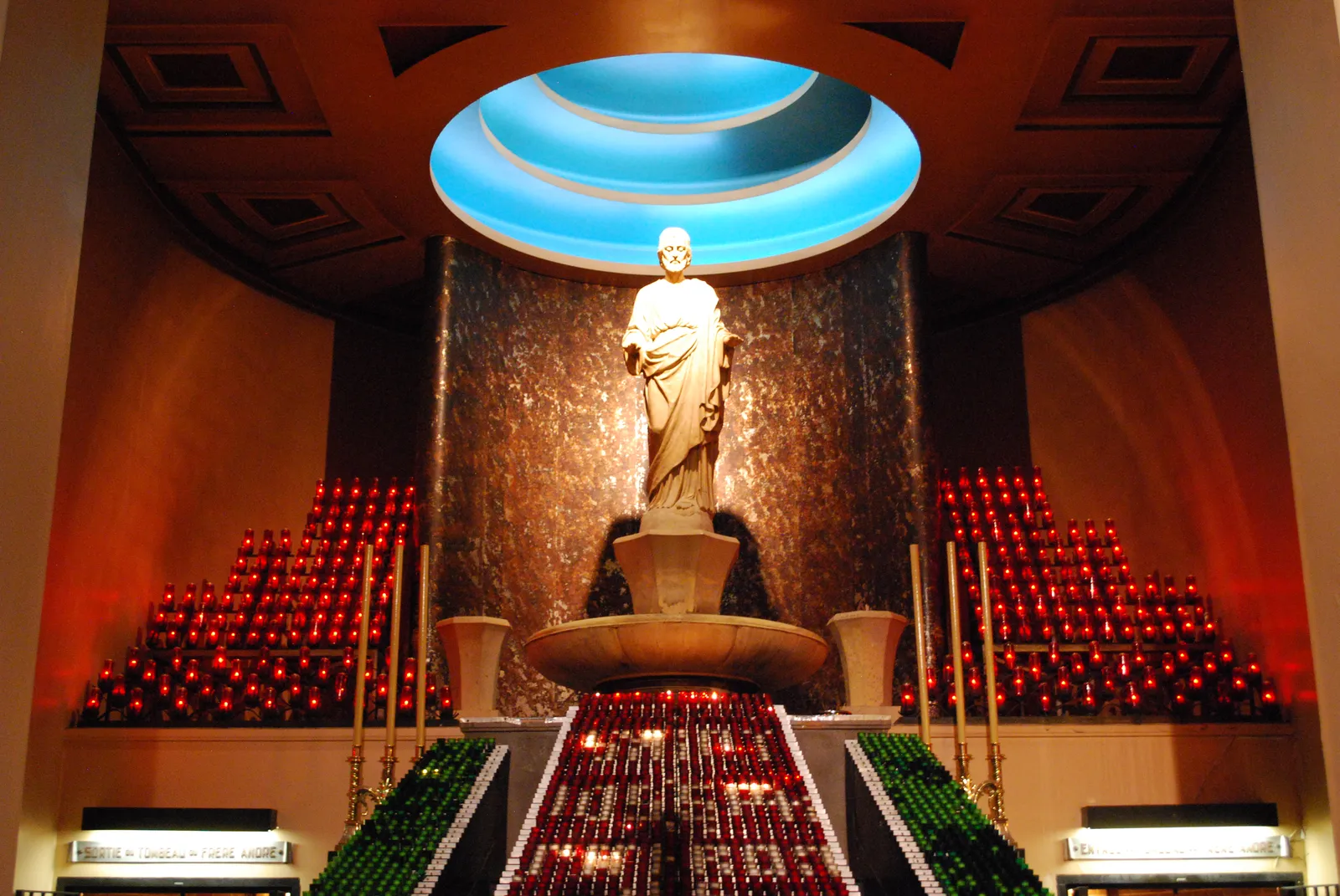 The votive chapel at Saint Joseph's Oratory in Montreal, banked tiers of red and green candles rising on both sides of a pale stone statue beneath a circular blue-lit ceiling opening