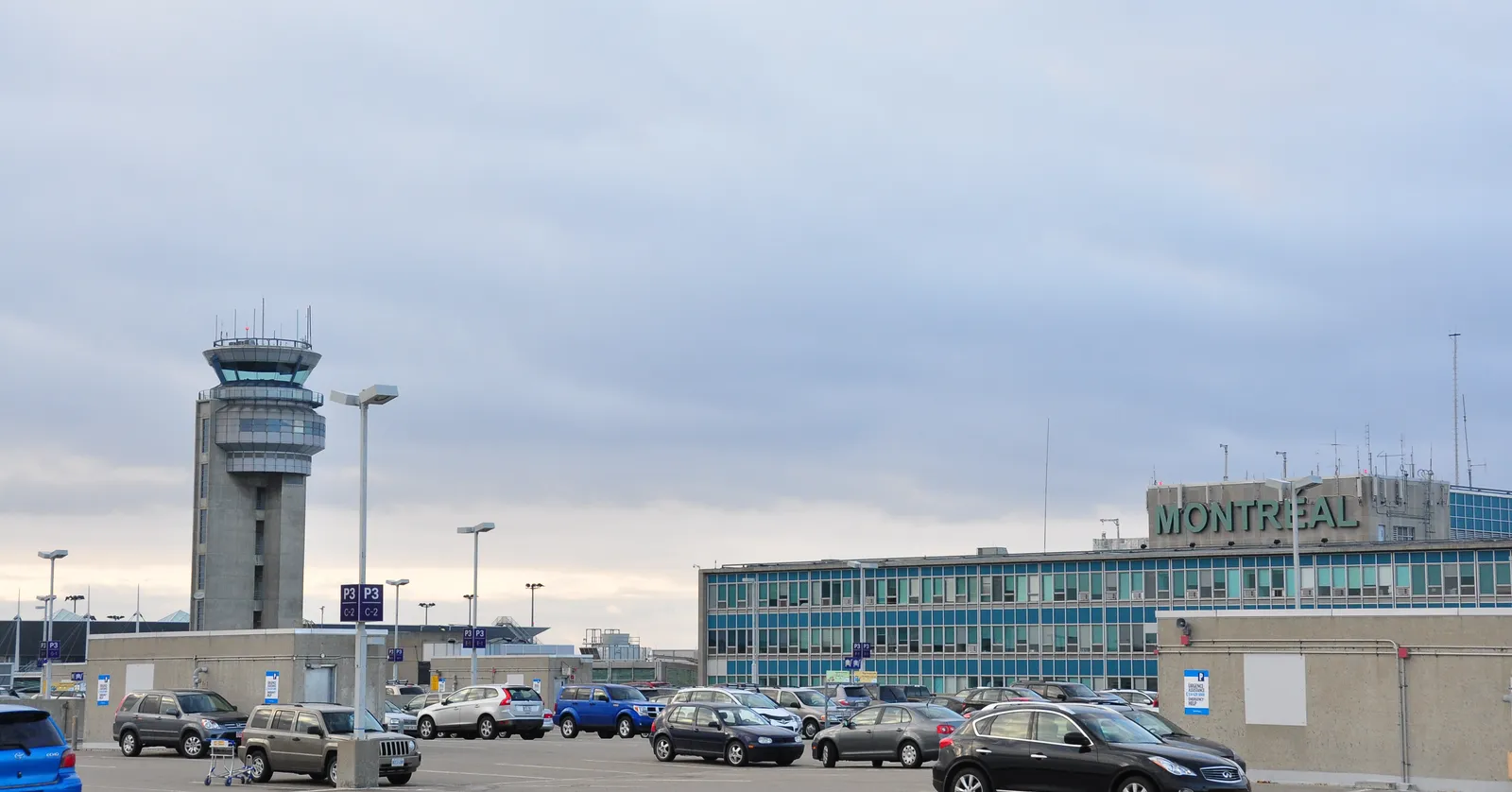 The front of the terminal at Montreal-Trudeau airport with MONTRÉAL spelled across its facade, the striped air traffic control tower standing beside it and cars parked along the kerb
