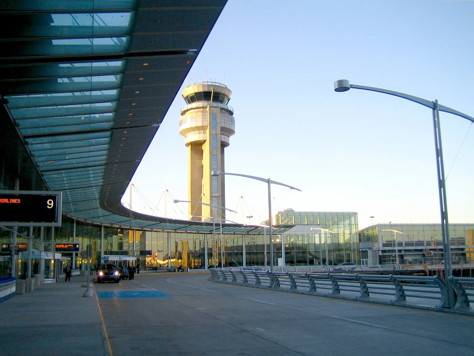 The United States departures kerbside at Montreal-Trudeau airport, a glass canopy running over door 9 with the striped air traffic control tower rising behind the terminal