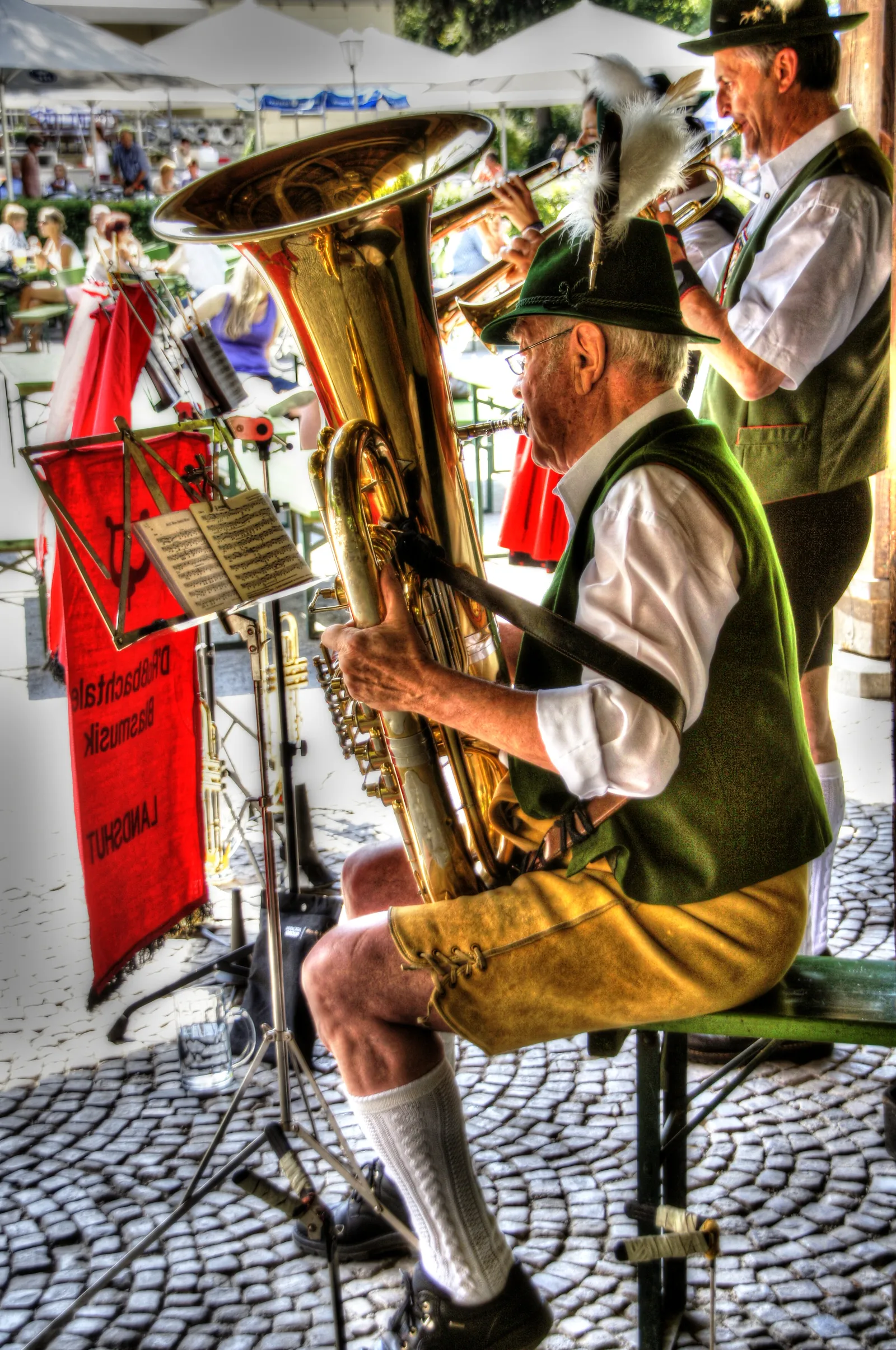 A Bavarian brass band musician in traditional lederhosen playing a tuba at the beer garden beneath the Chinesischer Turm, sheet music on a stand in front of him and a red banner reading Landshut behind