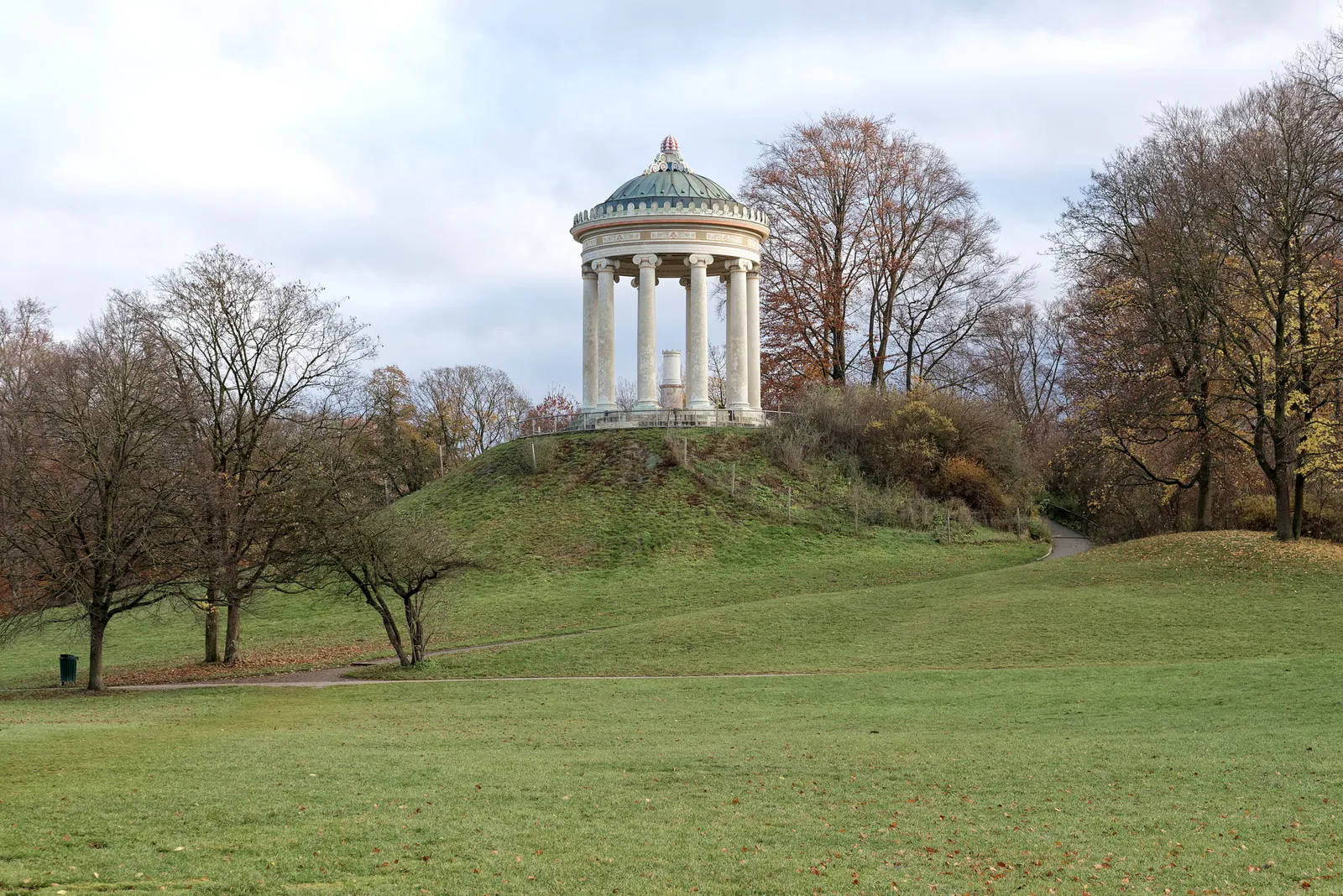 The Monopteros, a small round Greek-style temple with white columns and a green dome, standing on a grassy hill in Munich's Englischer Garten