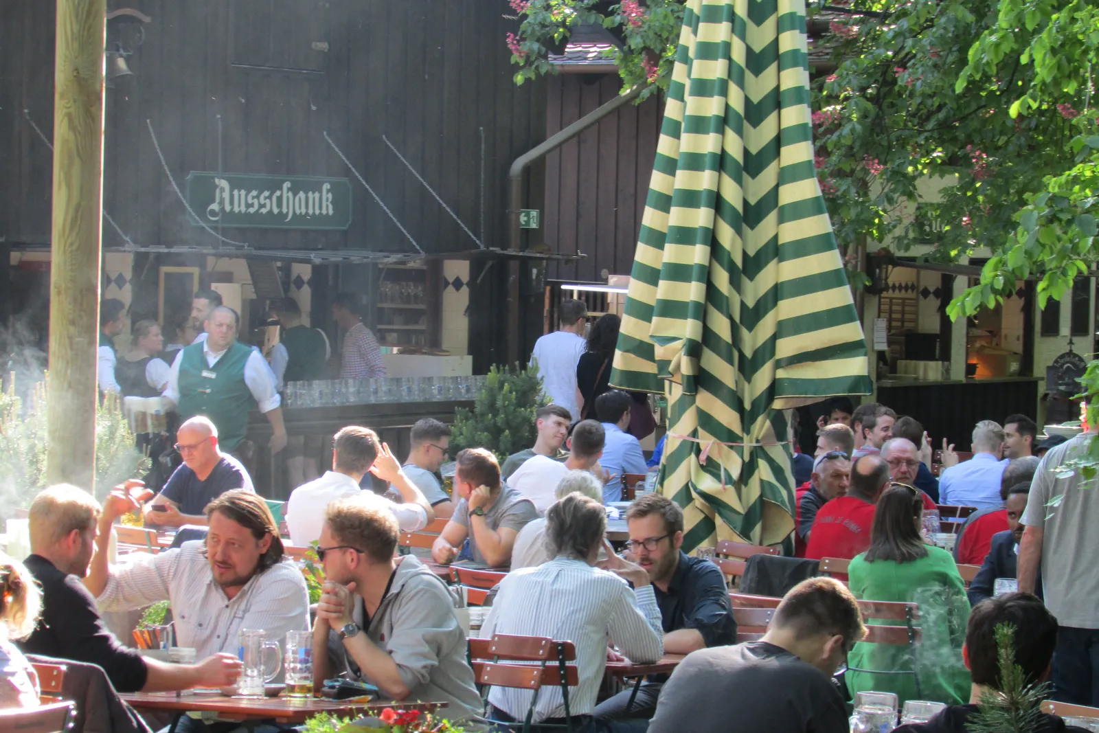 The beer garden at the Augustiner-Keller in Munich, with drinkers at wooden benches under chestnut trees and staff in green aprons working the self-service counter under the Ausschank sign