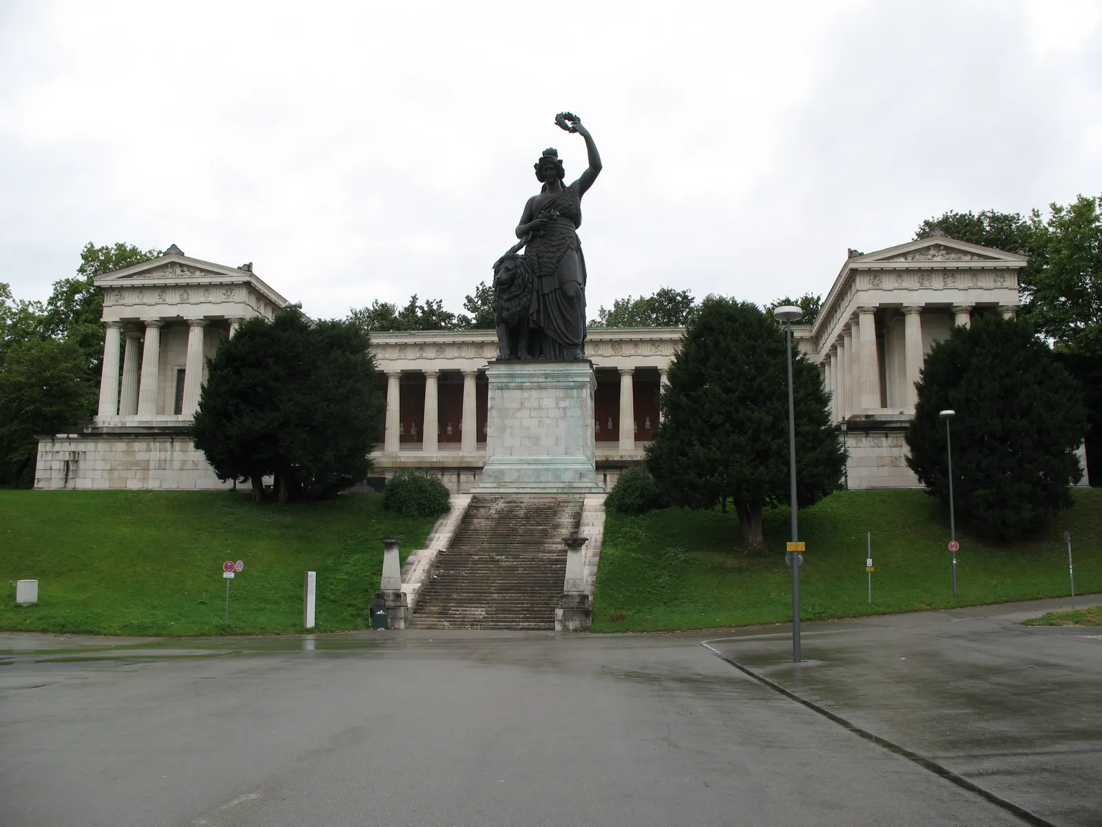 The bronze Bavaria statue on her plinth above the Theresienwiese in Munich, one arm raised, framed by the colonnaded wings of the Ruhmeshalle behind her