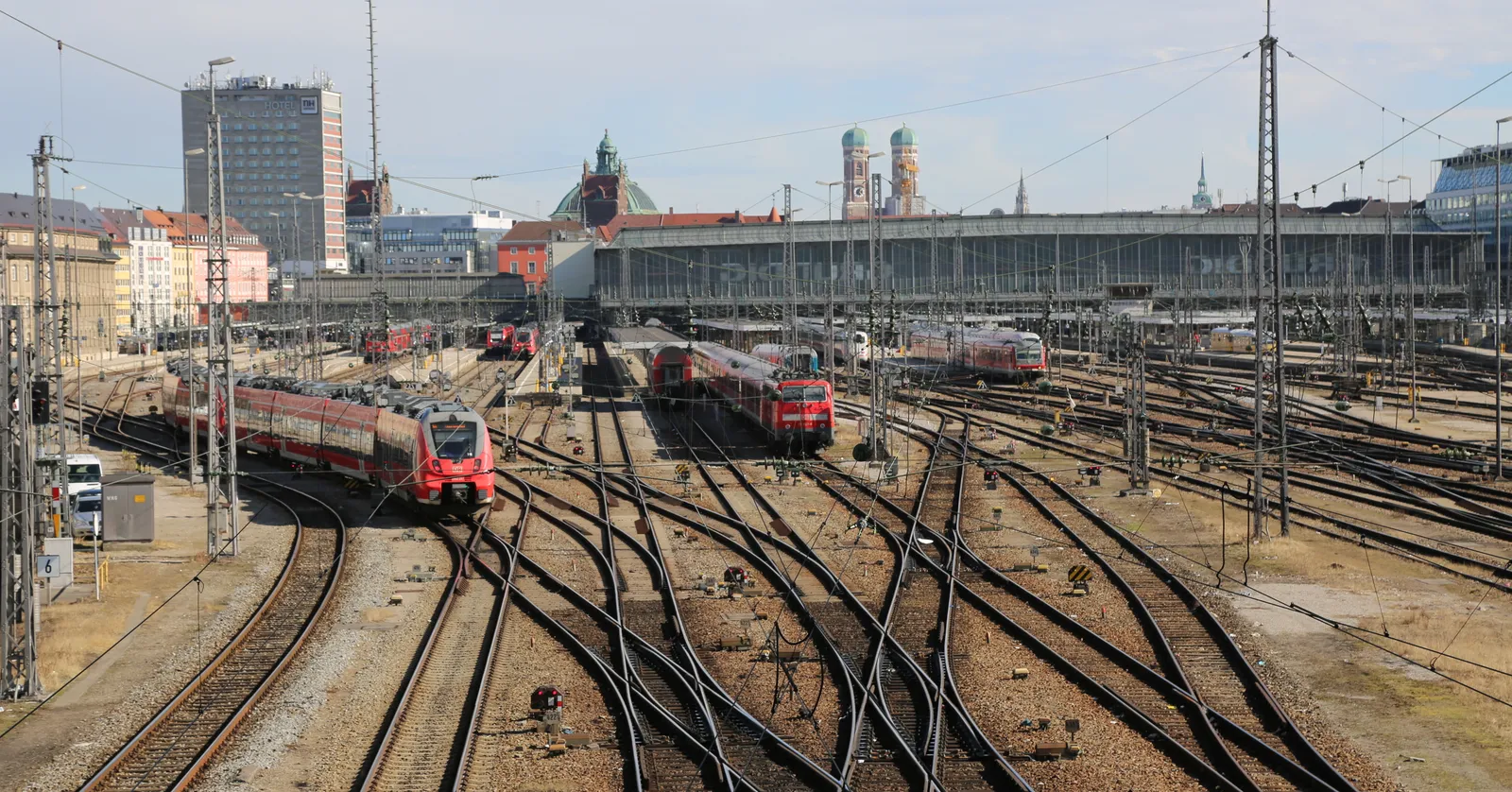 The track approach into Munich Hauptbahnhof seen from the Hackerbrücke, red regional trains standing under the platform canopies with the twin domes of the Frauenkirche on the skyline behind