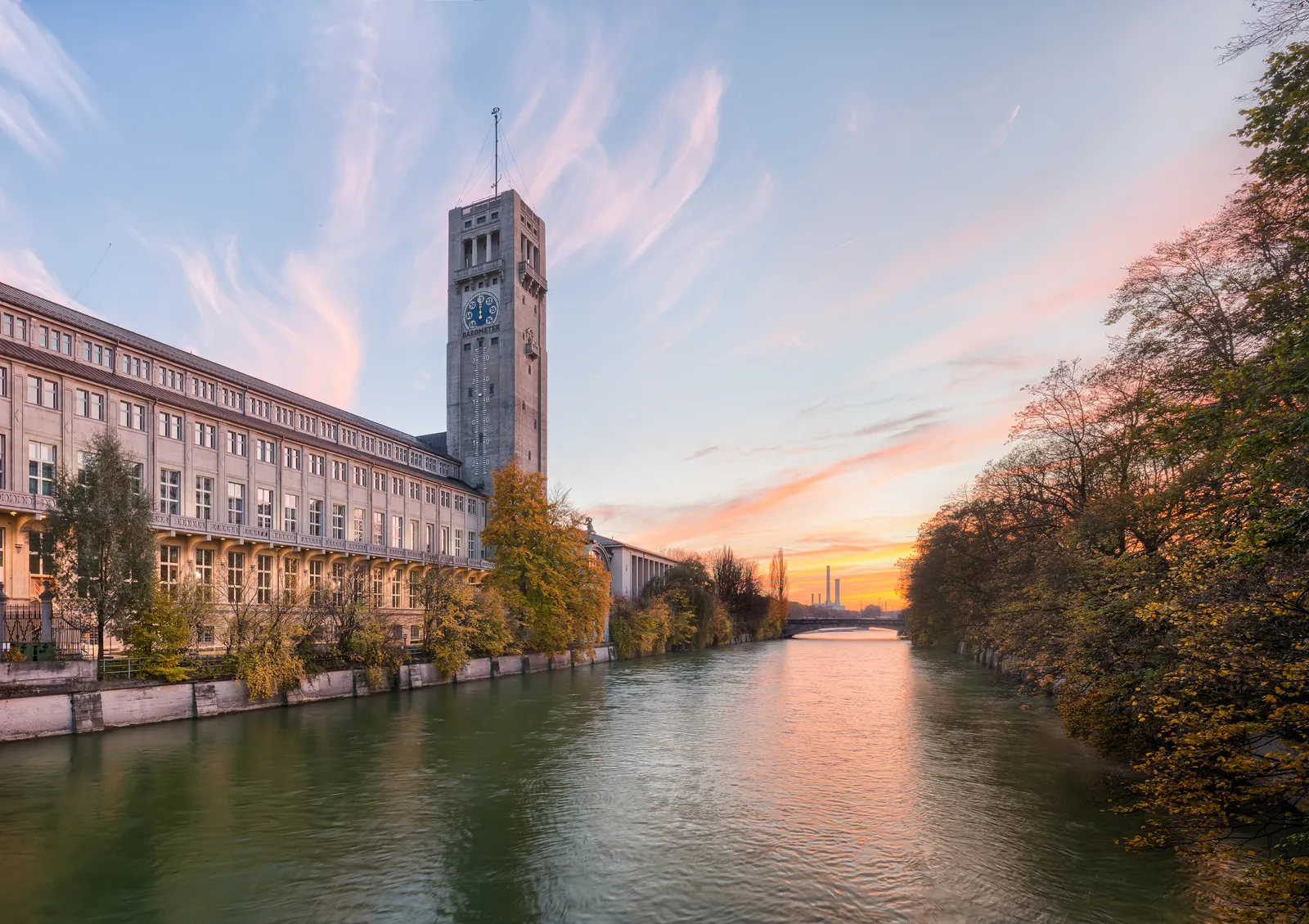 The Deutsches Museum's long riverside building and clock tower on Museum Island in Munich, seen along the Isar at sunset with the water reflecting pink and orange sky
