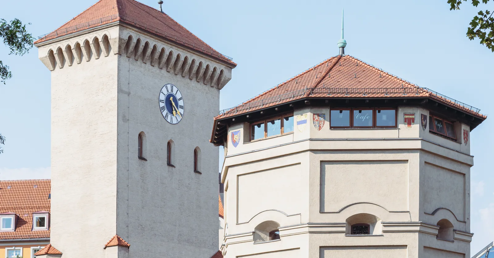 The two towers of Munich's Isartor gate against a clear blue sky, the tall central clock tower on the left and an octagonal side tower with painted heraldic shields and a cafe sign on the right