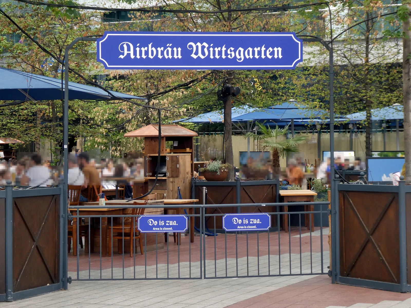 The gated entrance to the Airbräu Wirtsgarten beer garden at Munich Airport, a blue sign reading Airbräu Wirtsgarten over the gate, tables and blue umbrellas visible behind it