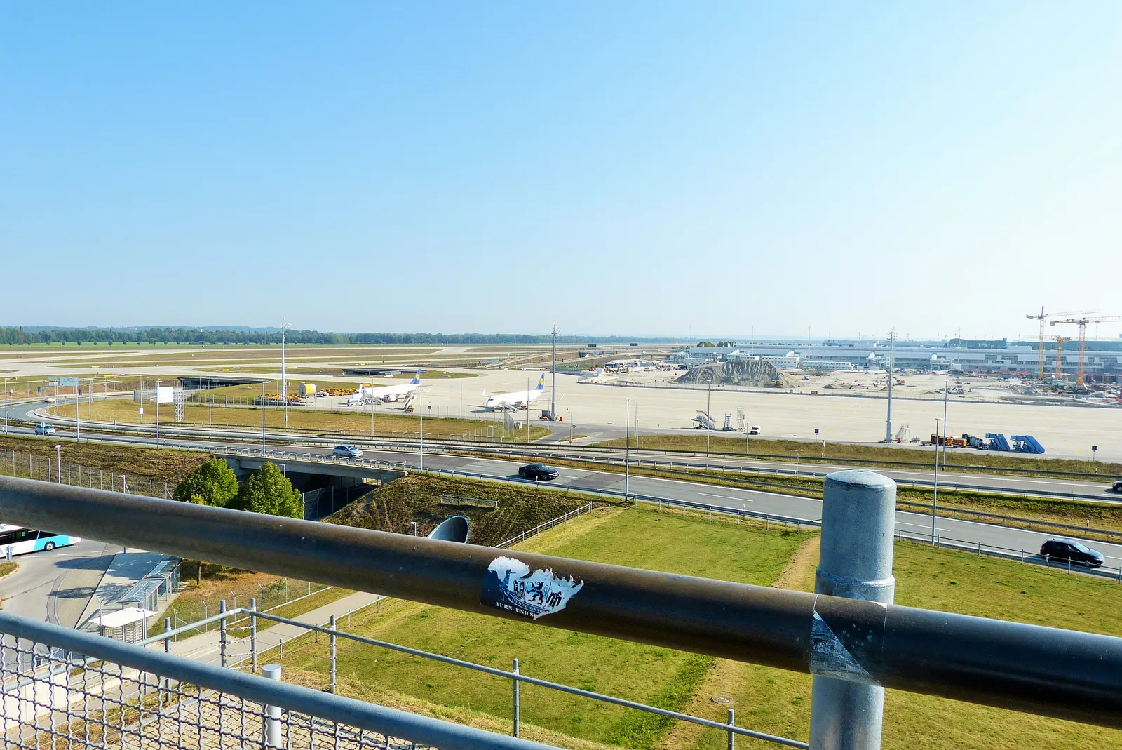 The railed viewing platform at the top of the Besucherhügel at Munich Airport, looking down over a taxiway and access road toward the terminal buildings and control tower in the distance