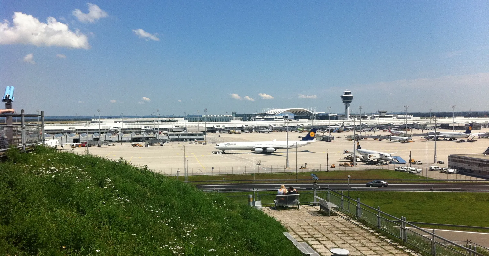Two people sitting on a bench on the grassy Besucherhügel at Munich Airport, coin-operated binoculars beside them, looking out over the apron at a Lufthansa jet and the control tower under a blue sky