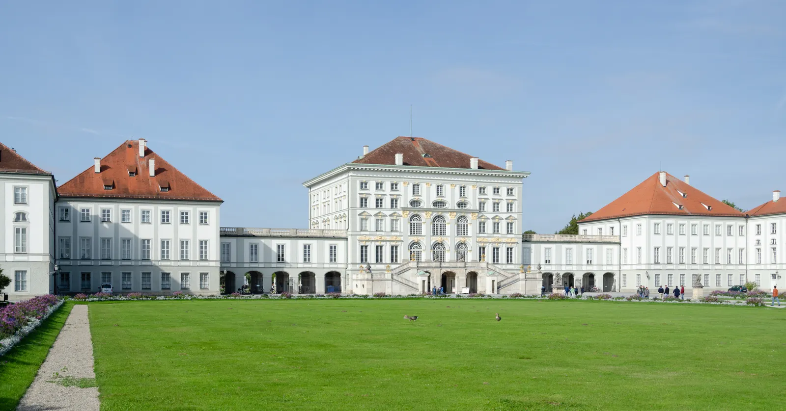 The wide white baroque facade of Schloss Nymphenburg with its red-tiled roofs and curving colonnaded wings, seen across the open lawn in front of it under a clear blue sky