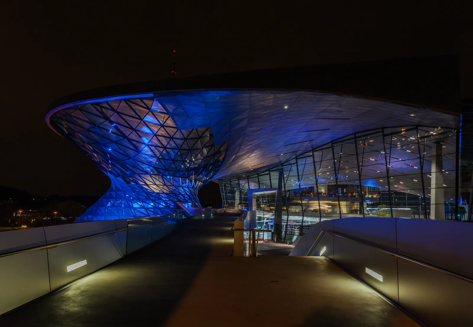 The curved, funnel-shaped glass and steel roof of BMW Welt in Munich lit blue at night, with a lit walkway ramp leading up into the building