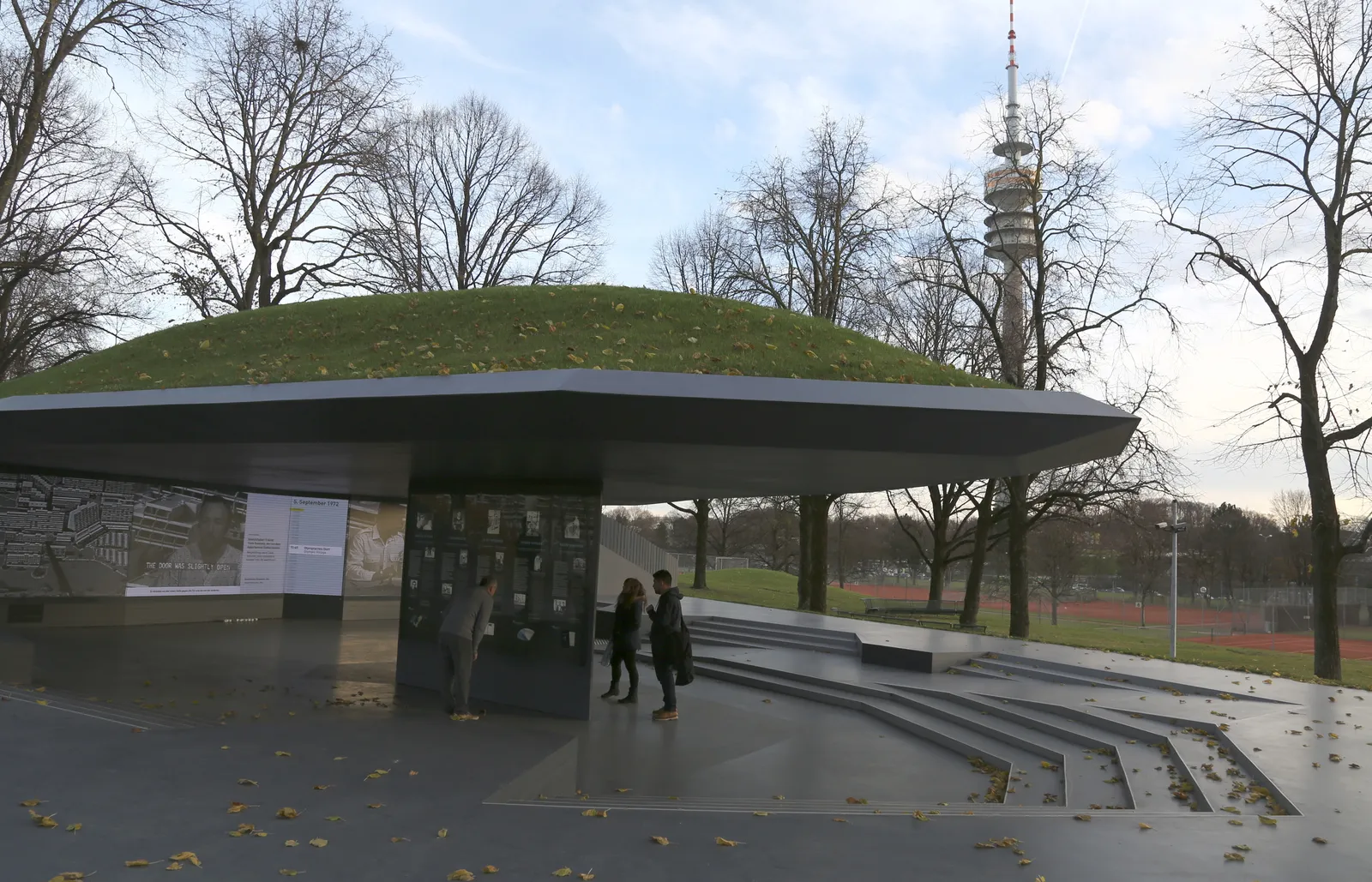 The low, grass-roofed pavilion of the Erinnerungsort Olympia-Attentat memorial in Munich's Olympiapark, visitors reading information panels at its entrance with the Olympiaturm visible behind