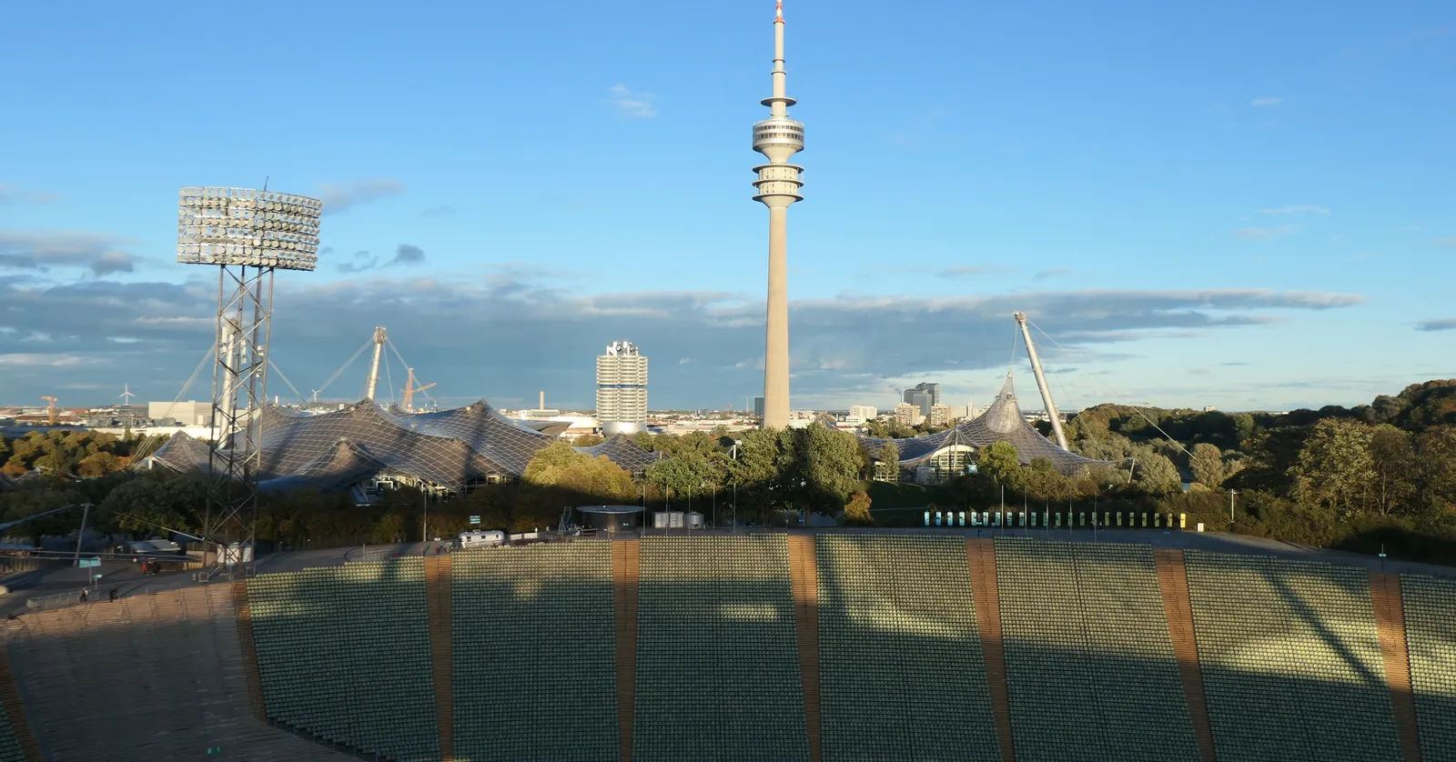 The Olympiapark's tent-roof stadium seen from the stands, with the Olympiaturm television tower and the four-cylinder BMW headquarters tower rising behind it under a clear blue sky