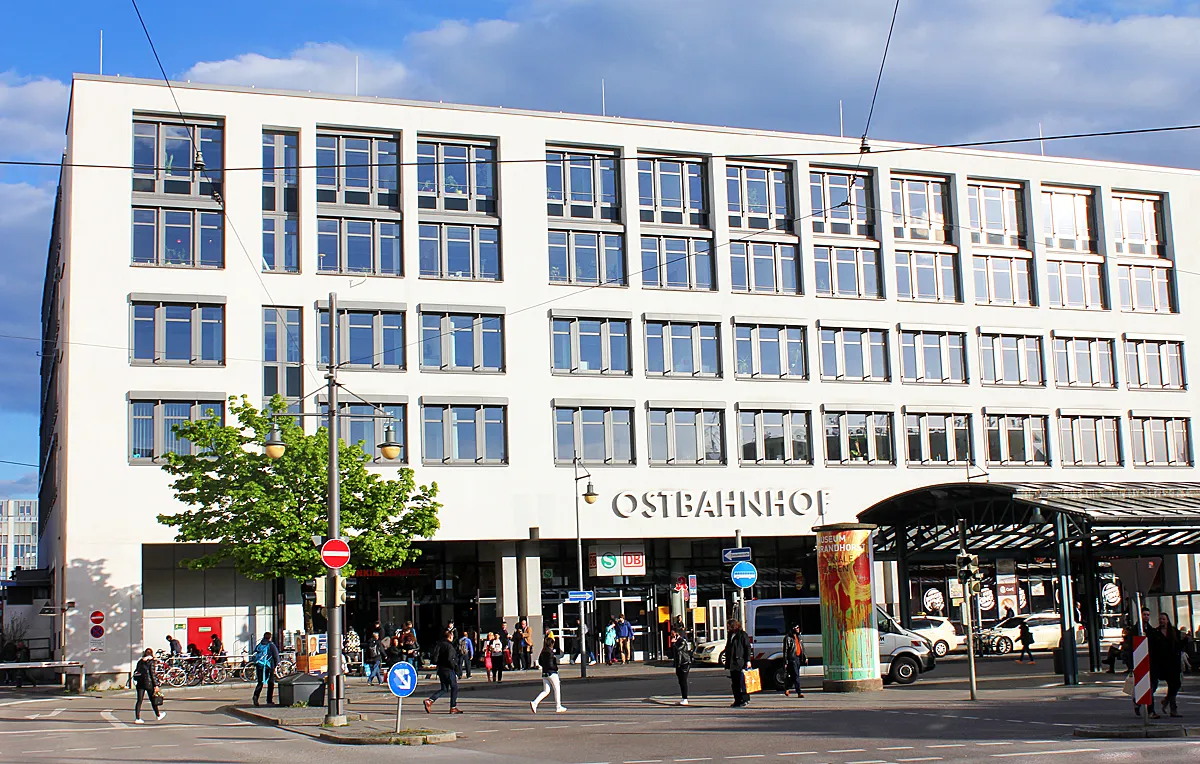 The white facade of Munich Ostbahnhof with OSTBAHNHOF spelled out in large letters above the entrance, trees and pedestrians on the street in front