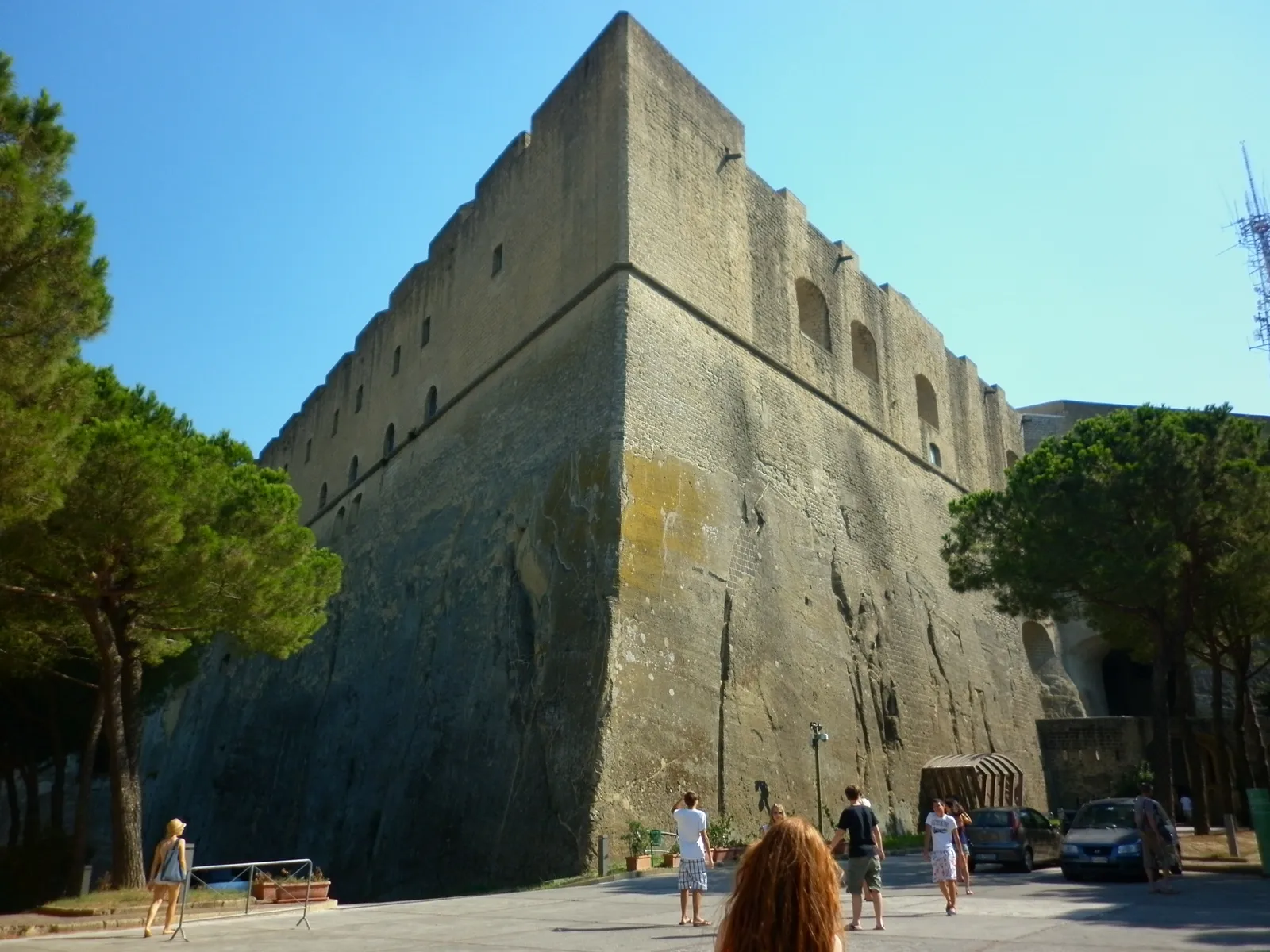The stone ramparts of Castel Sant'Elmo in Naples rising in a sharp angled point above a pine-shaded terrace, with people walking along the wall at its base