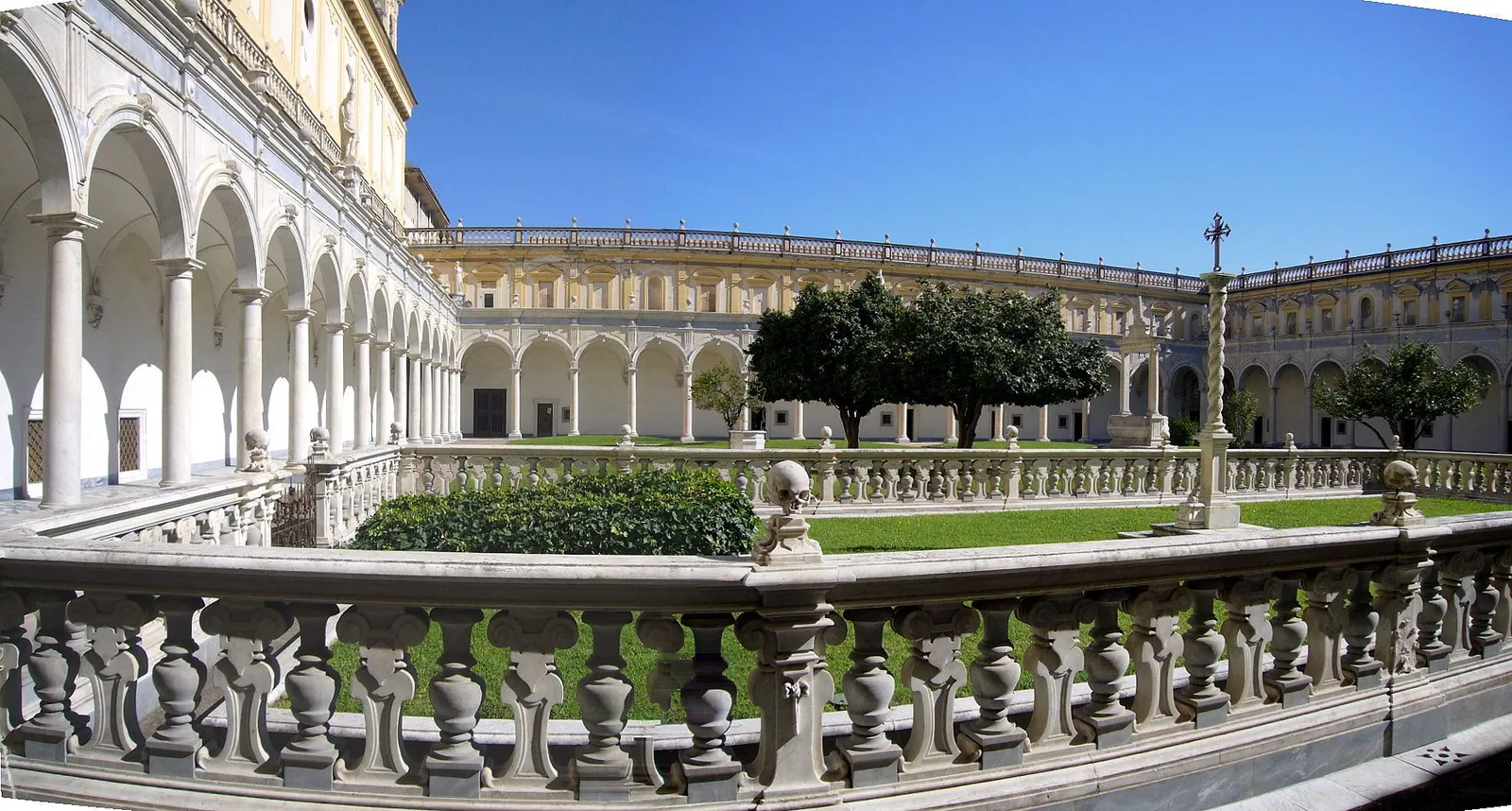 The Great Cloister of the Certosa di San Martino, a white arcaded courtyard with a lawn and trees, and a marble balustrade in the foreground topped with carved skulls