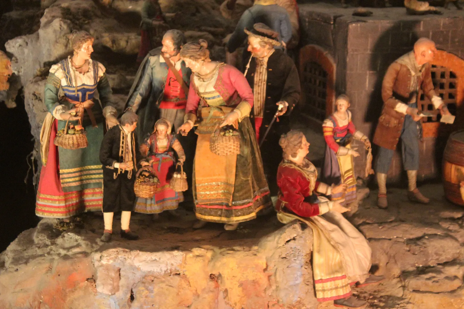 A group of eighteenth-century terracotta figures in silk costume from the Cuciniello nativity scene in Naples, standing on a modelled rock face beside a cellar door