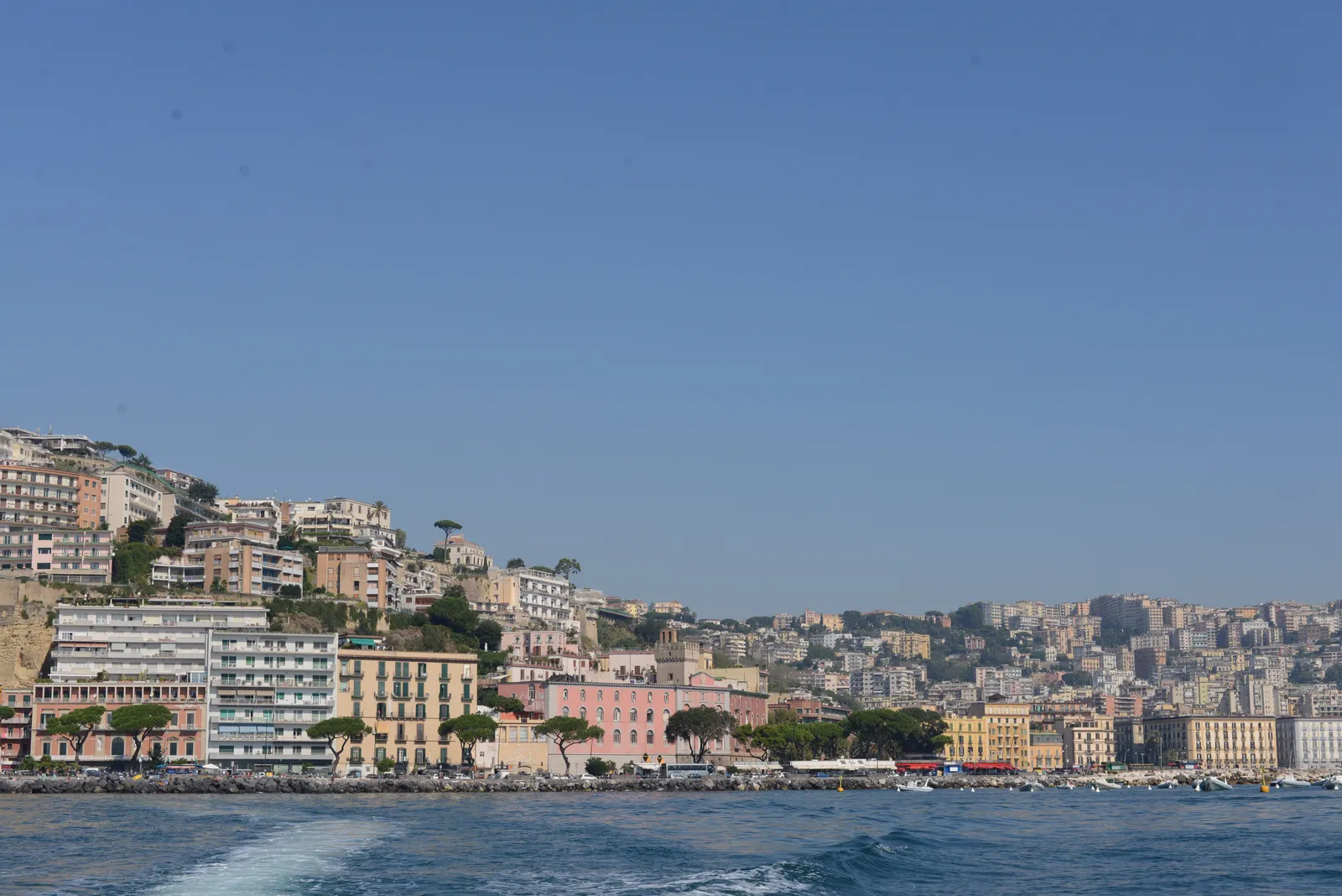 The Mergellina waterfront in Naples seen from the water, pastel apartment blocks stacked up the green Posillipo slope above a line of quayside buildings