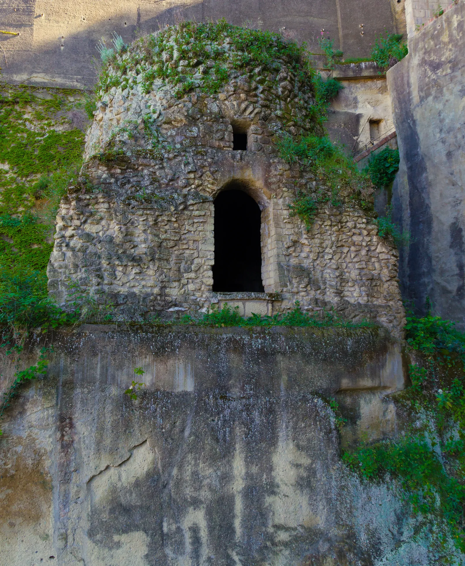 The Roman tomb known as Virgil's, a rough stone burial chamber with a single arched doorway, set into a cliff face and overgrown with grass and creepers