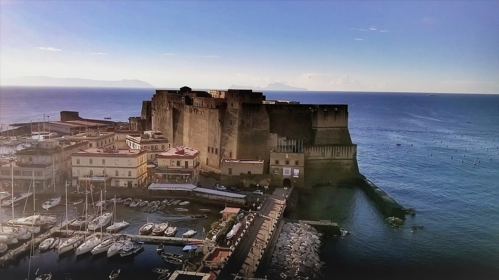 Castel dell'Ovo in Naples, a broad tufa fortress filling a small island, with the yacht harbour and low ochre houses of Borgo Marinari packed along its landward side