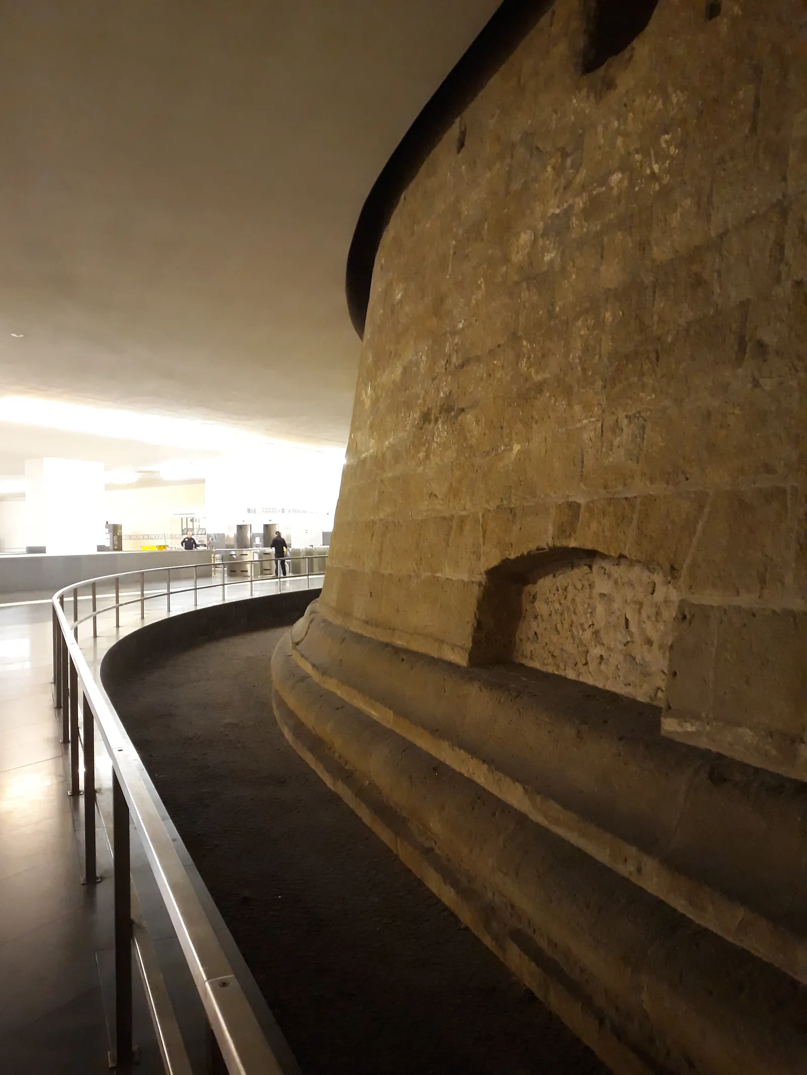 The curved stone base of a medieval defensive tower rising through the concourse of Municipio metro station in Naples, with a walkway and railing curving around it