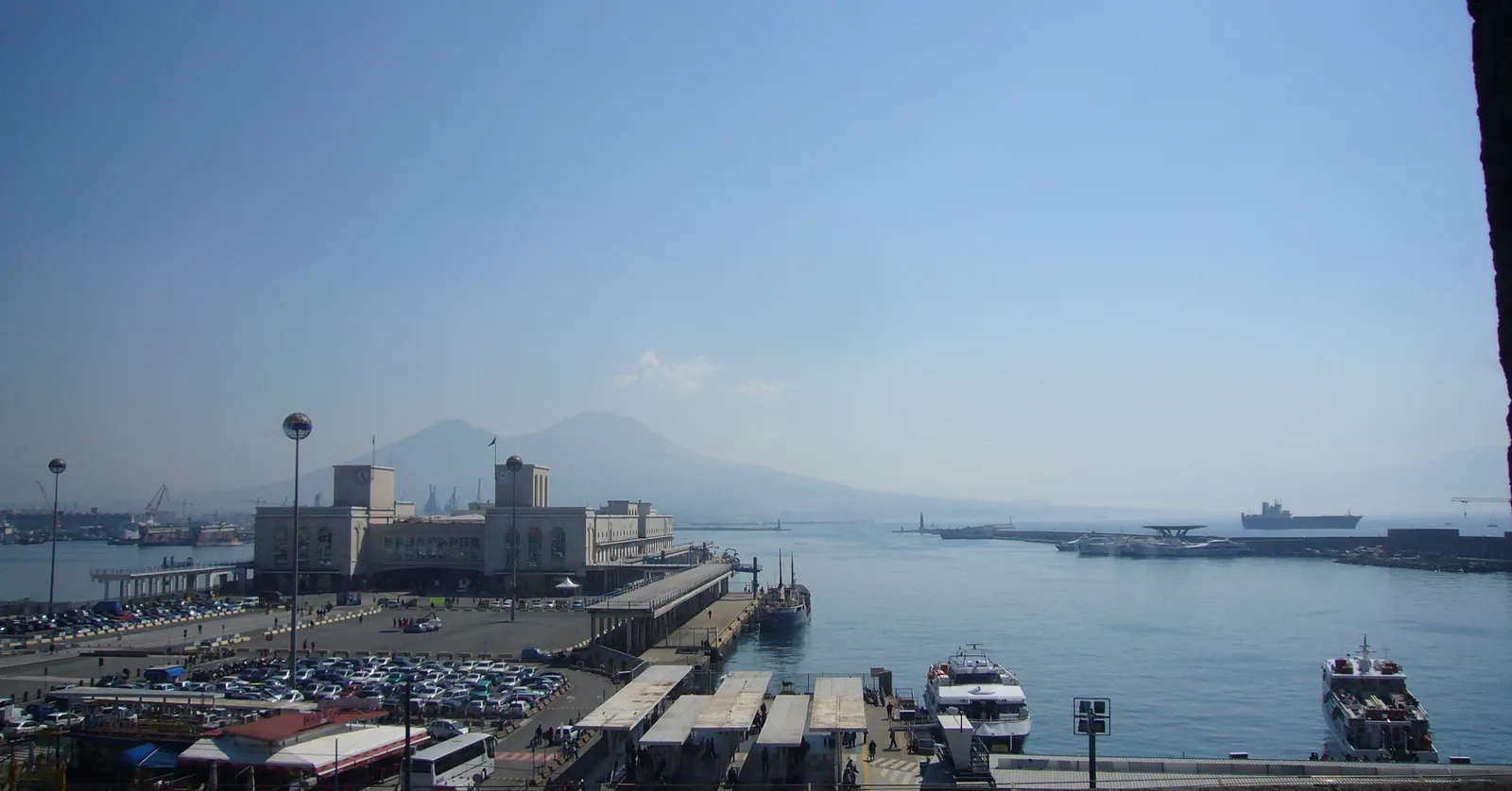 The port of Naples seen from above, with ferries and hydrofoils along the quays, the long pale terminal buildings behind them and Vesuvius across the bay