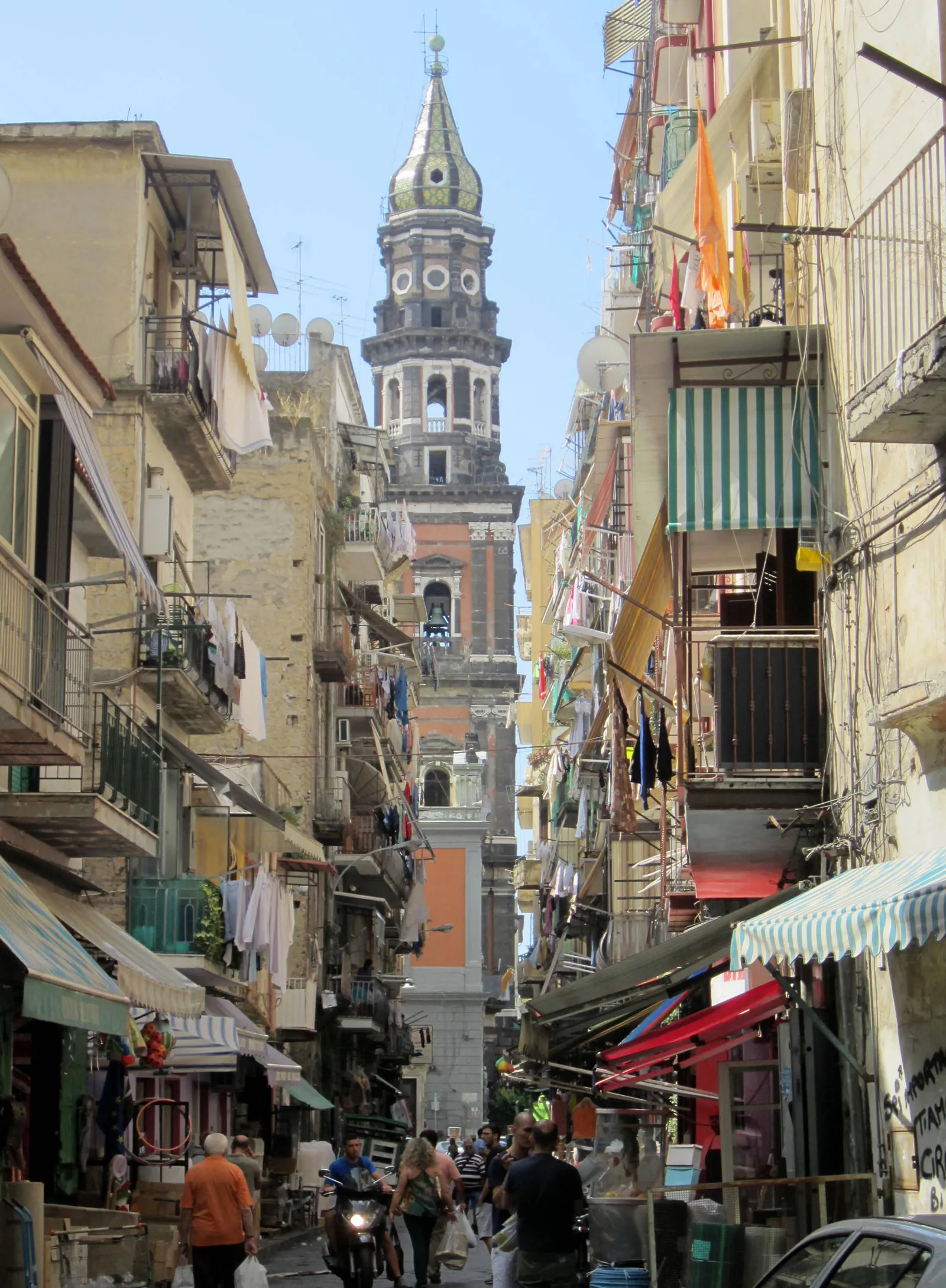 The bell tower of Santa Maria del Carmine in Naples seen from a narrow street hung with laundry, its tiled spire rising above the rooftops