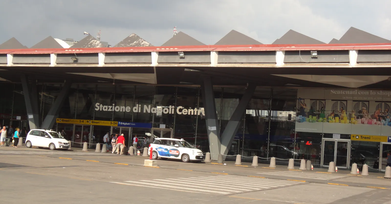 The front of Napoli Centrale station, a low glass and concrete building with a zigzag roof and the words Stazione di Napoli Centrale above the entrance doors