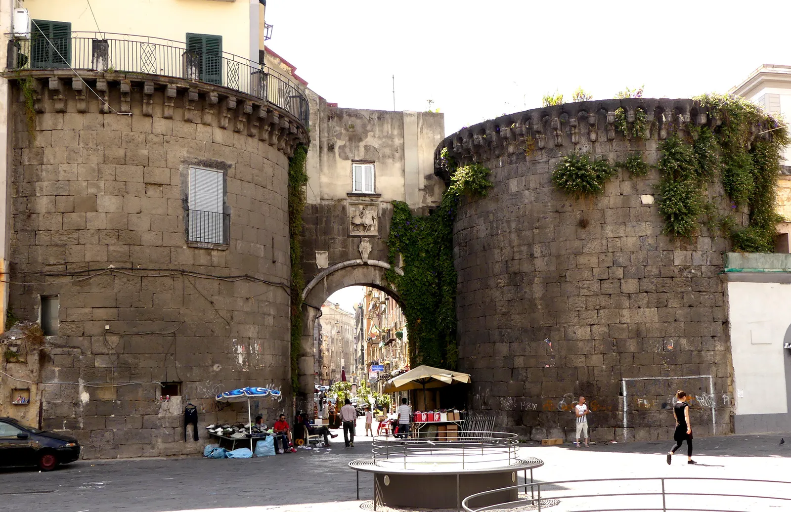 The Porta Nolana gate in Naples, a stone arch between two squat round towers, with market stalls and shoppers visible through the opening