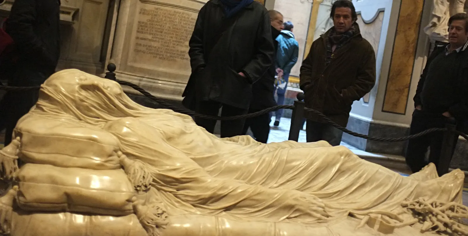 Giuseppe Sanmartino's Veiled Christ in the Sansevero Chapel in Naples, a life-size marble figure lying under a carved marble shroud, with visitors standing behind a rope at the far side