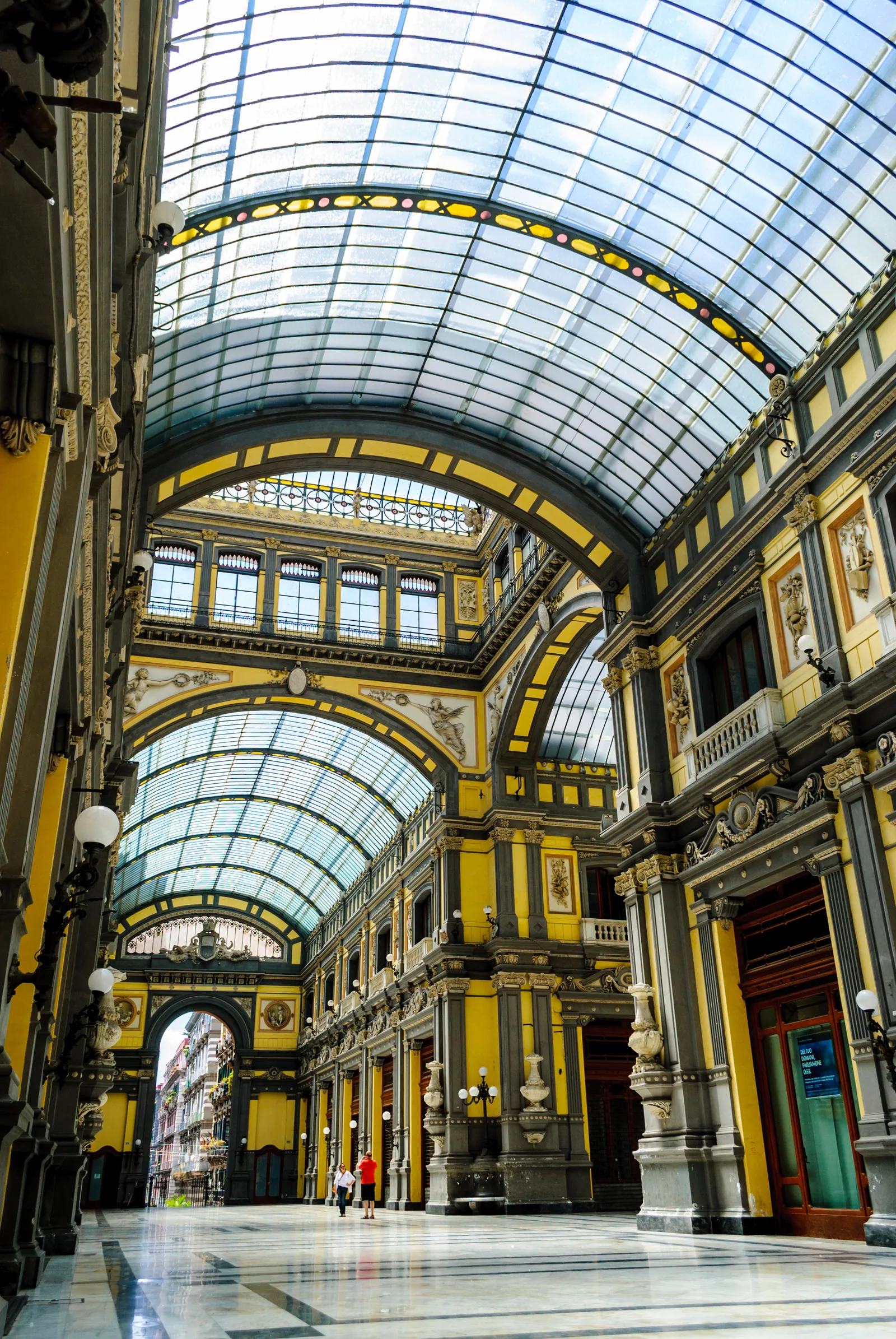 Inside the Galleria Principe di Napoli, a yellow and grey shopping arcade running under a long barrel vault of iron ribs and glass, with shopfronts and lamps along both sides