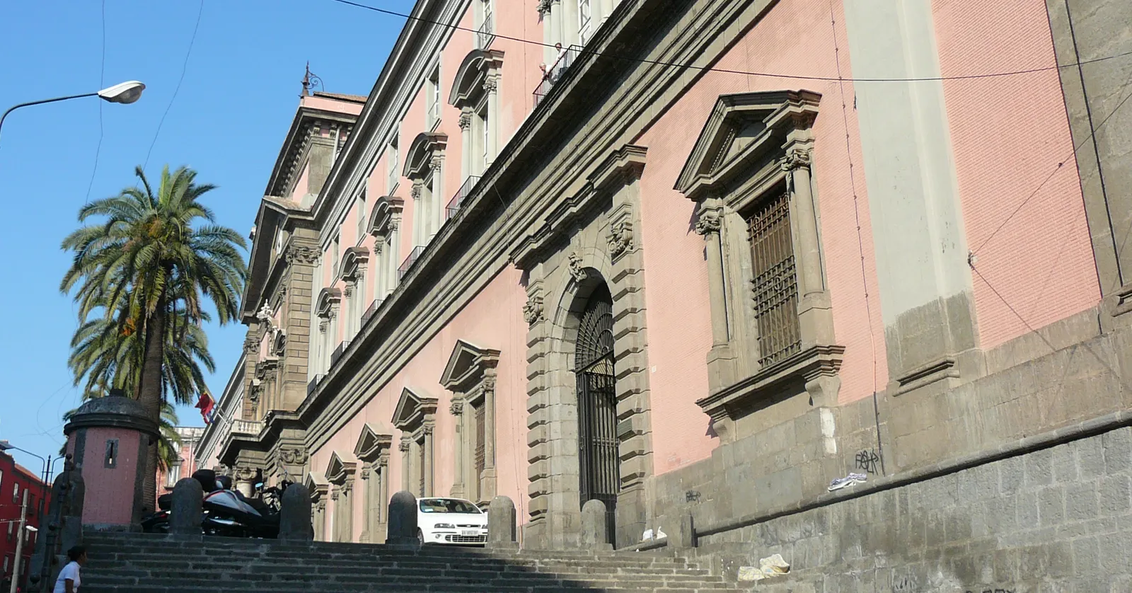 The long pink and grey stone front of the National Archaeological Museum in Naples, seen from the steps below it, with an arched doorway, barred ground-floor windows and a palm tree at the corner