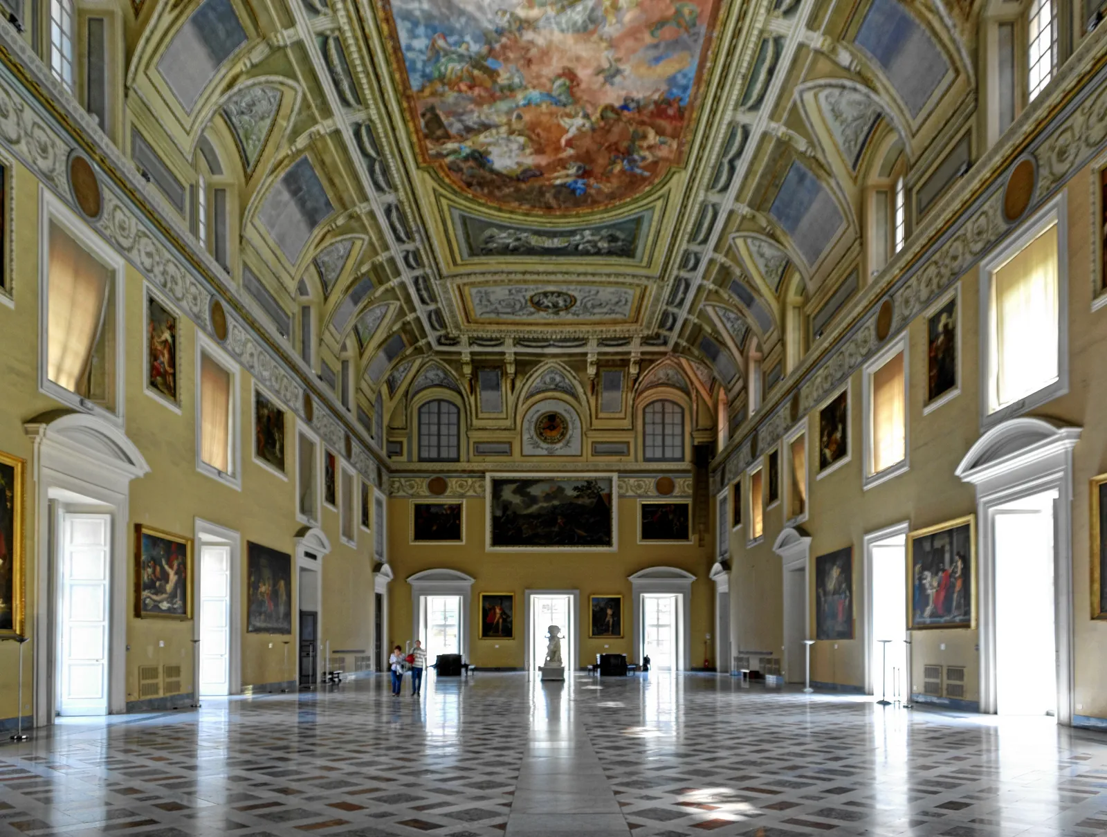 The Sundial Hall of the Naples archaeological museum, a long room with pale walls hung with paintings, tall windows down both sides and a single large fresco filling the vaulted ceiling
